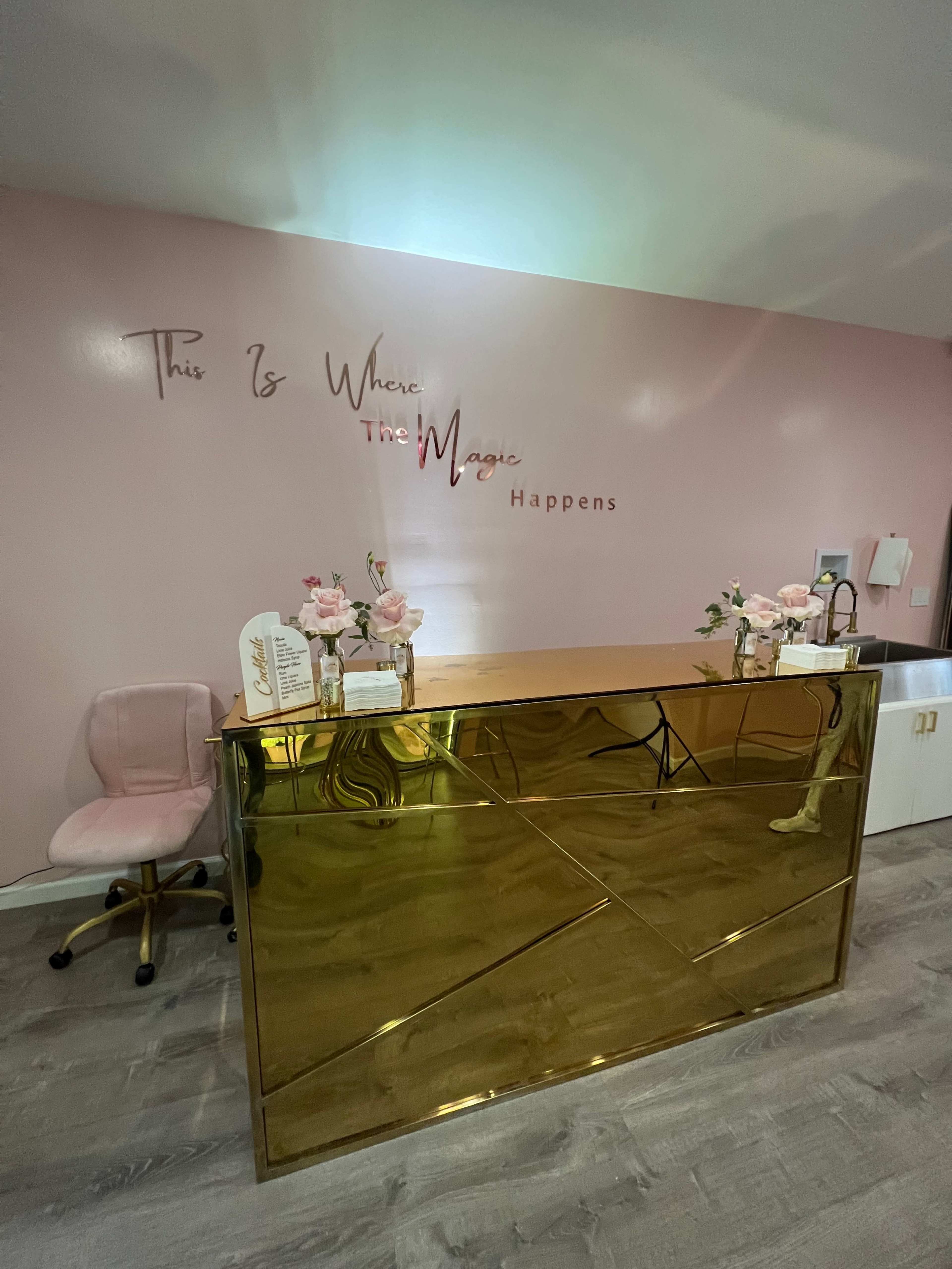 The image shows a reception area featuring a shiny gold desk and a pink accent wall with the phrase "This Is Where The Magic Happens" displayed.