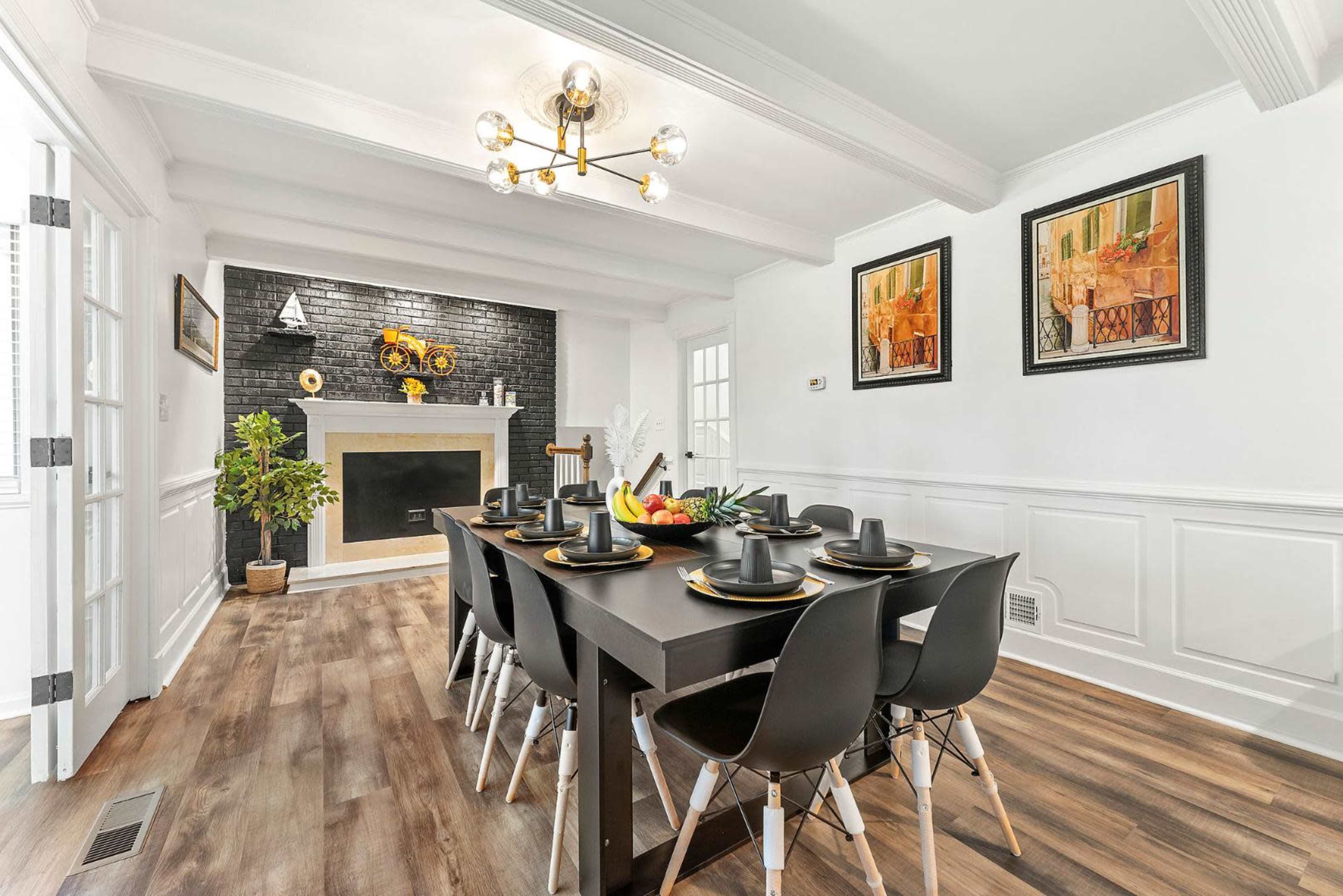 The image shows a modern dining room with a large table set for eight, featuring black chairs and decorative items, alongside a fireplace and framed artwork on the walls.