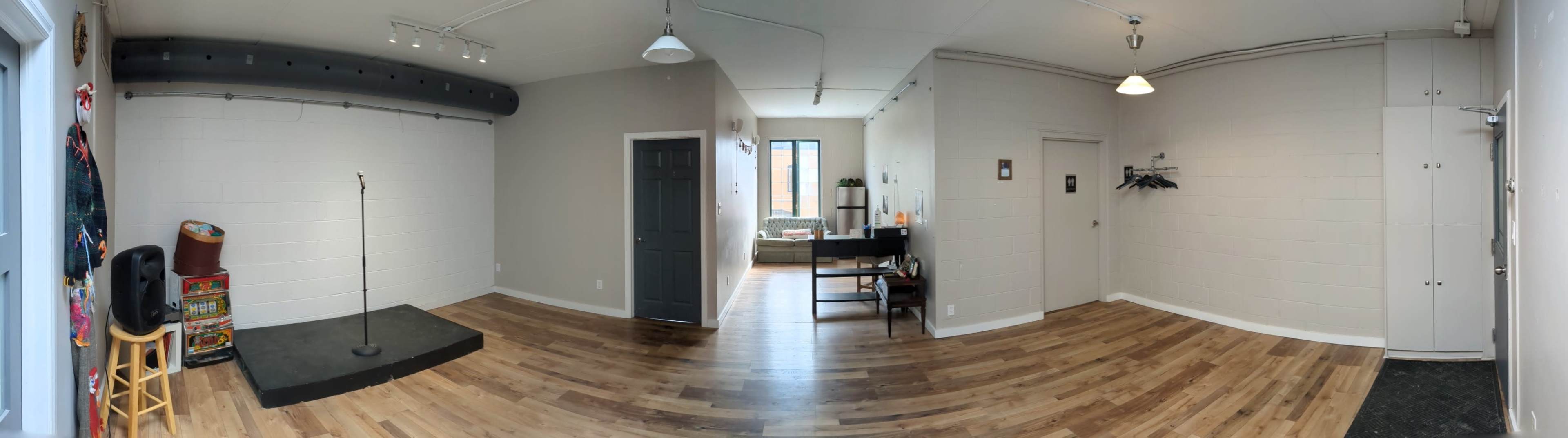 A spacious room with wooden flooring, a small stage with a microphone stand, a desk and chair, and a couch visible through a window.
