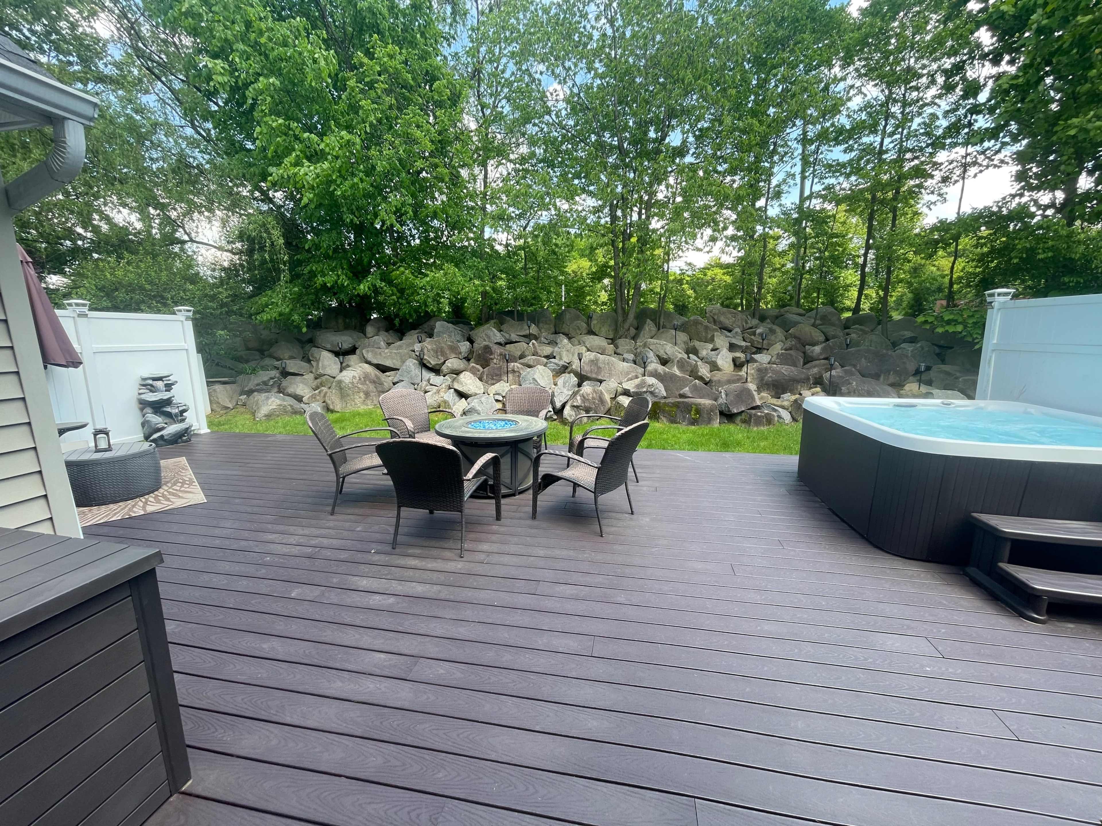 A wooden deck with a seating area featuring a table and chairs, alongside a hot tub and a backdrop of large rocks and greenery.