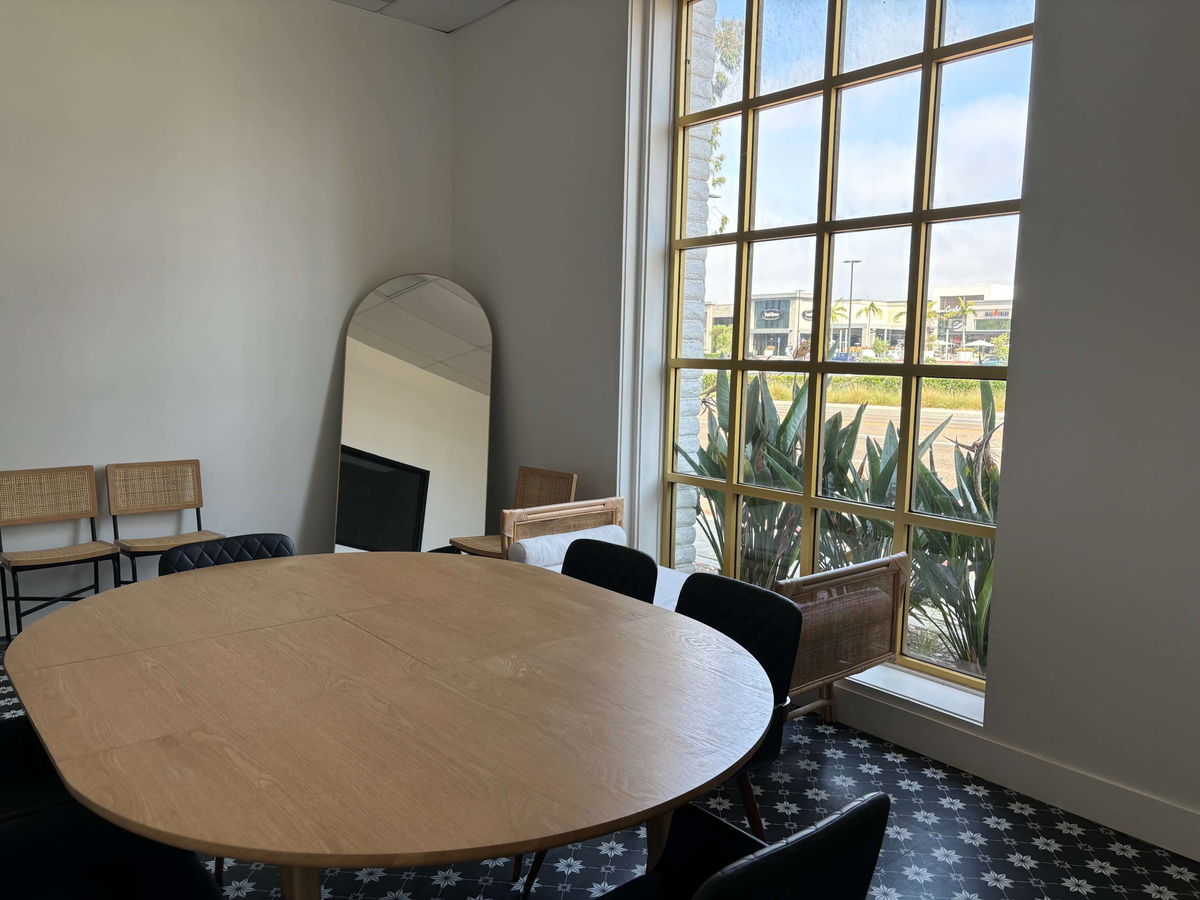 A round wooden table sits in a well-lit room near a large window with a mirror and seating arranged around it.