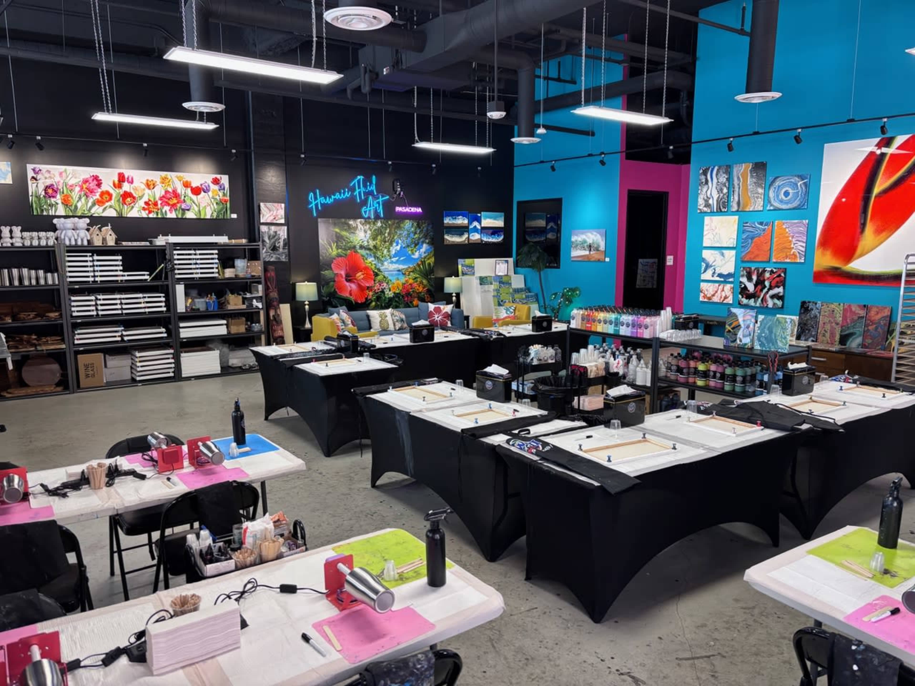The image shows an art studio set up for a painting class, featuring multiple tables with canvases, art supplies, and colorful wall decorations.