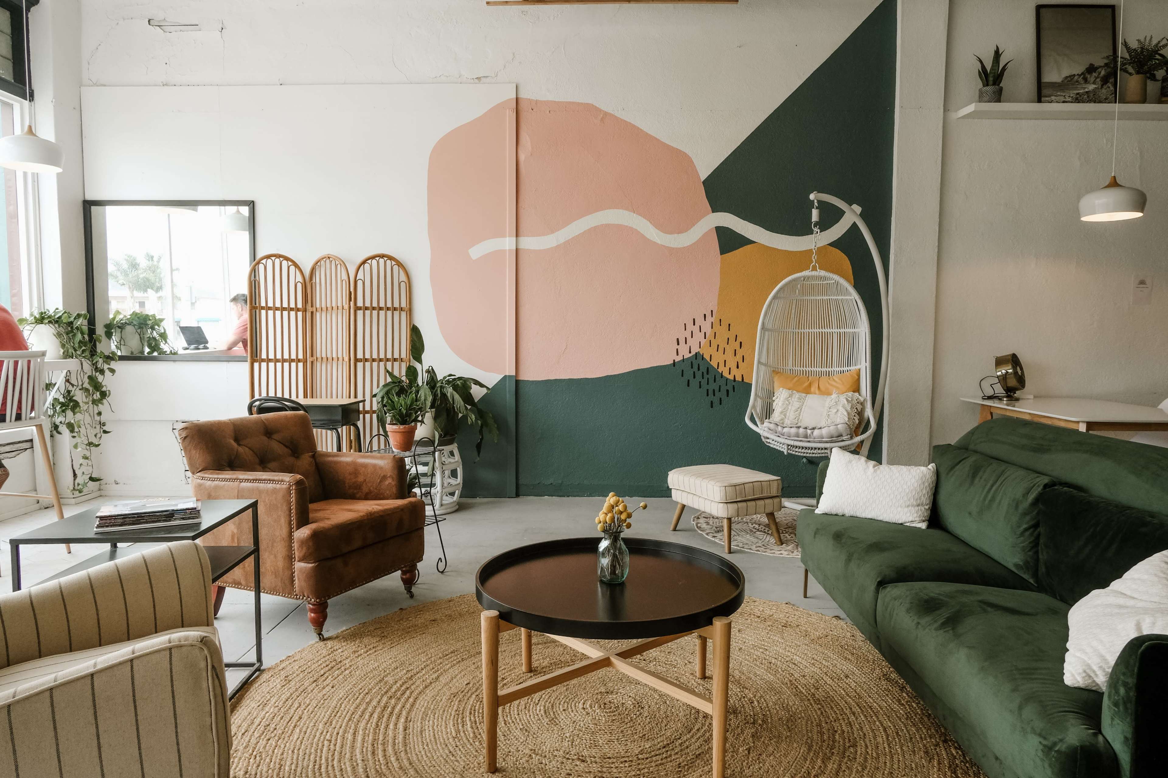 A modern, airy interior space with a green sofa, a hanging chair, a round table, and an artistic wall mural in soft colors.