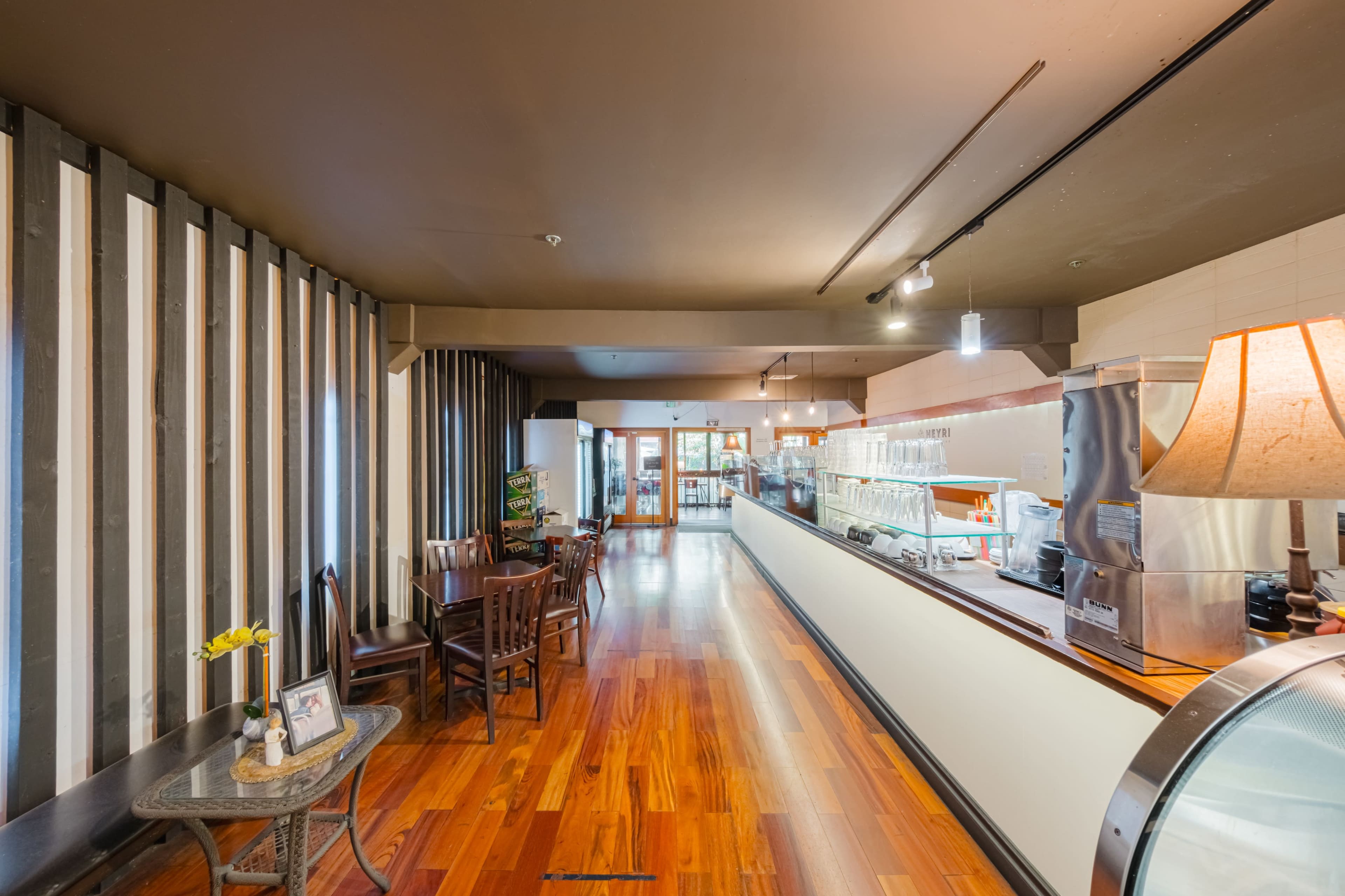 A modern café interior featuring wooden flooring, a counter with a beverage display, and several wooden chairs arranged throughout the space.