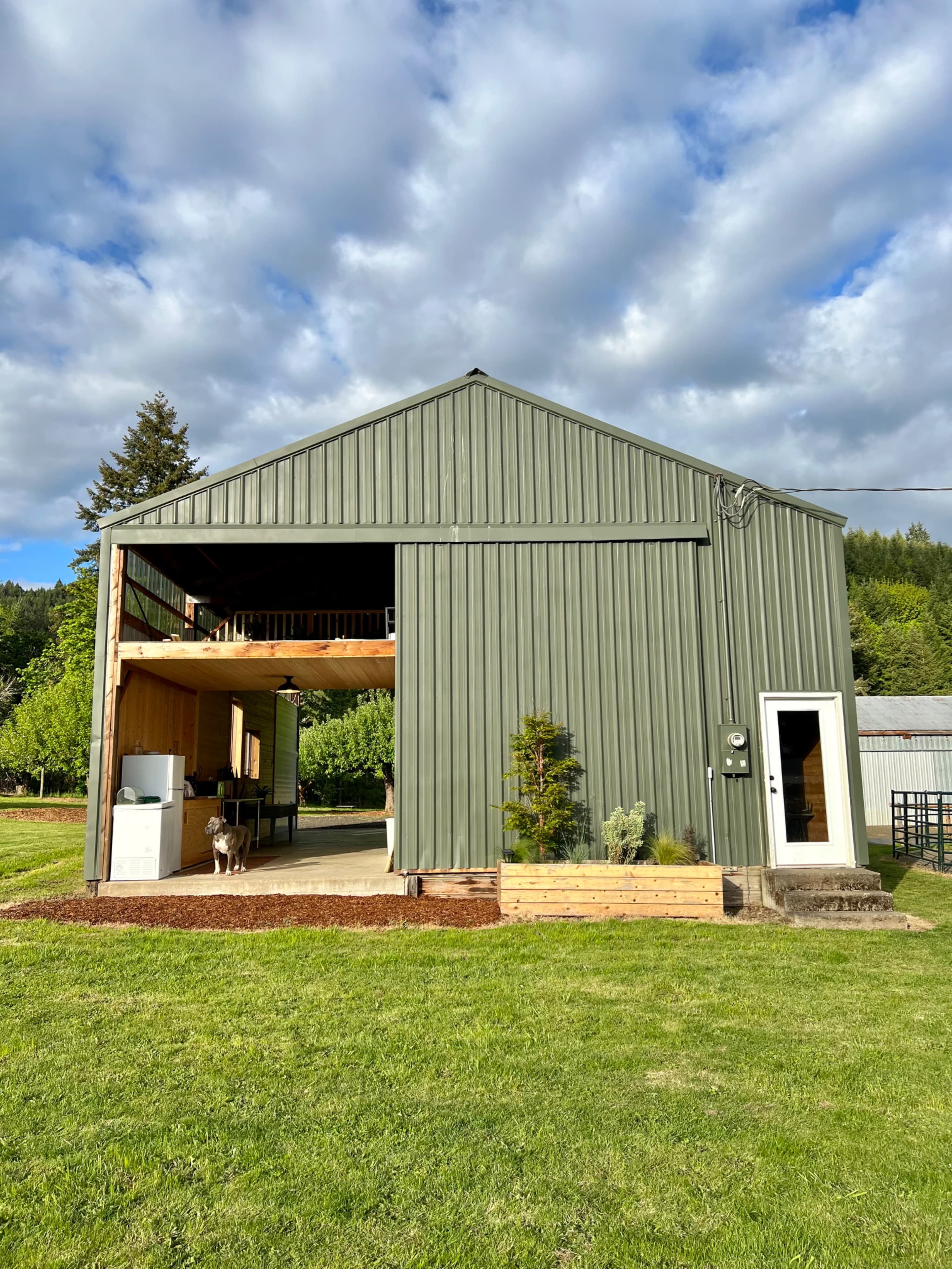 A large green metal barn features an open front with a wooden deck, set on a grassy landscape under a partly cloudy sky.