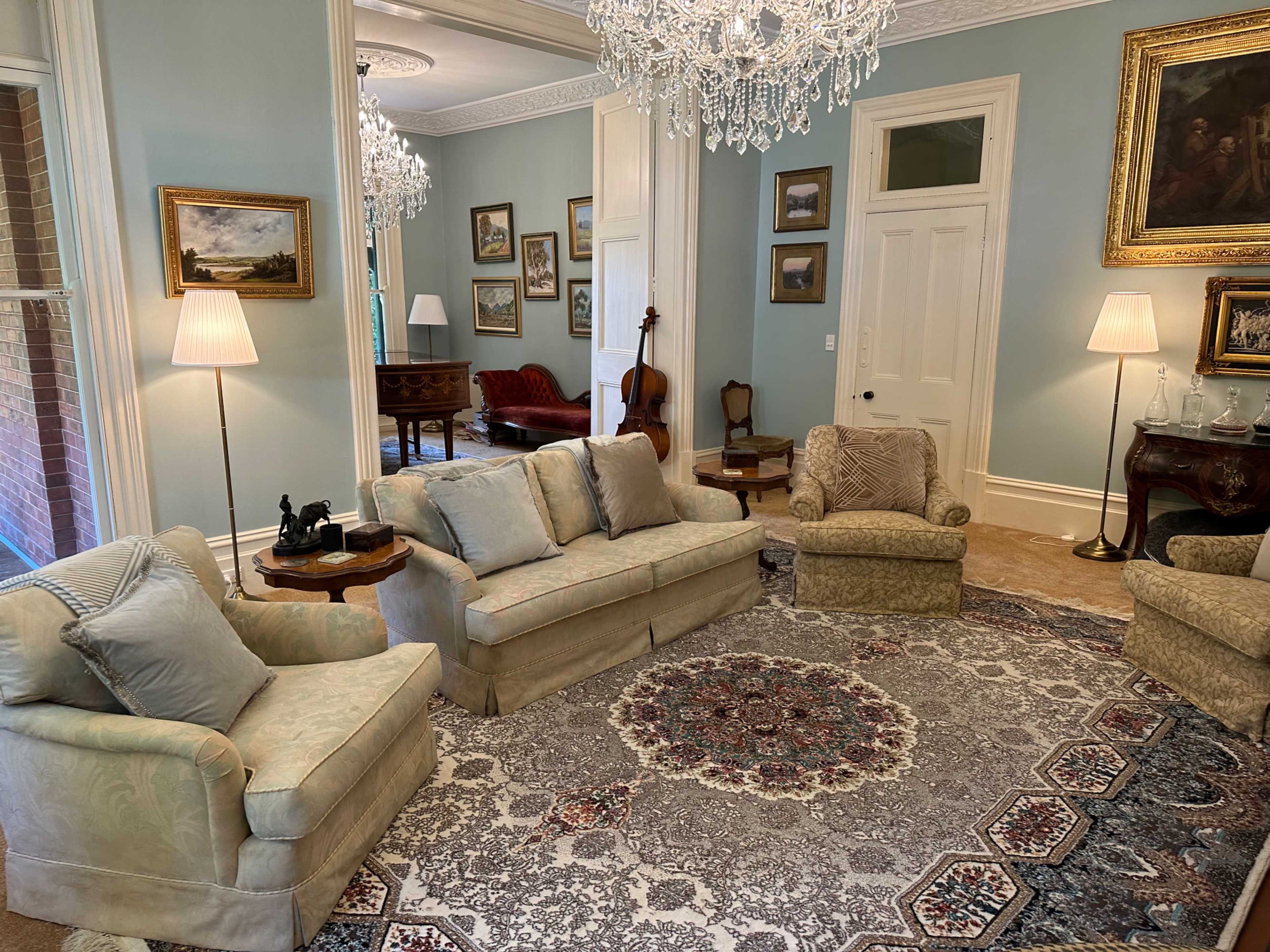 A cozy living room with a patterned rug, light-colored sofas, and a chandelier, featuring framed artwork and a wooden piano in the background.