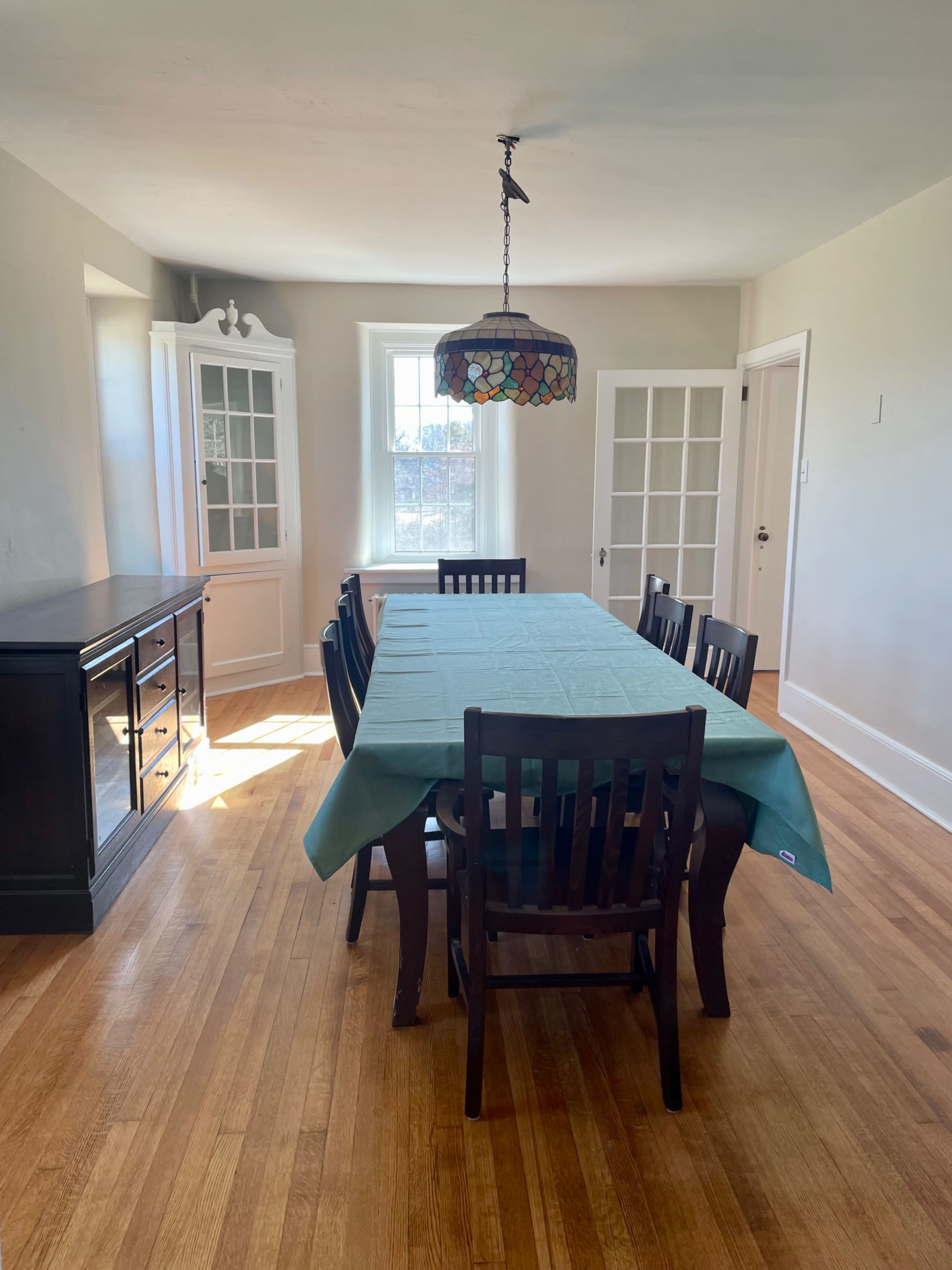 A dining room with a large, rectangular table covered by a green tablecloth, surrounded by chairs, and a stained glass pendant light hanging above it.
