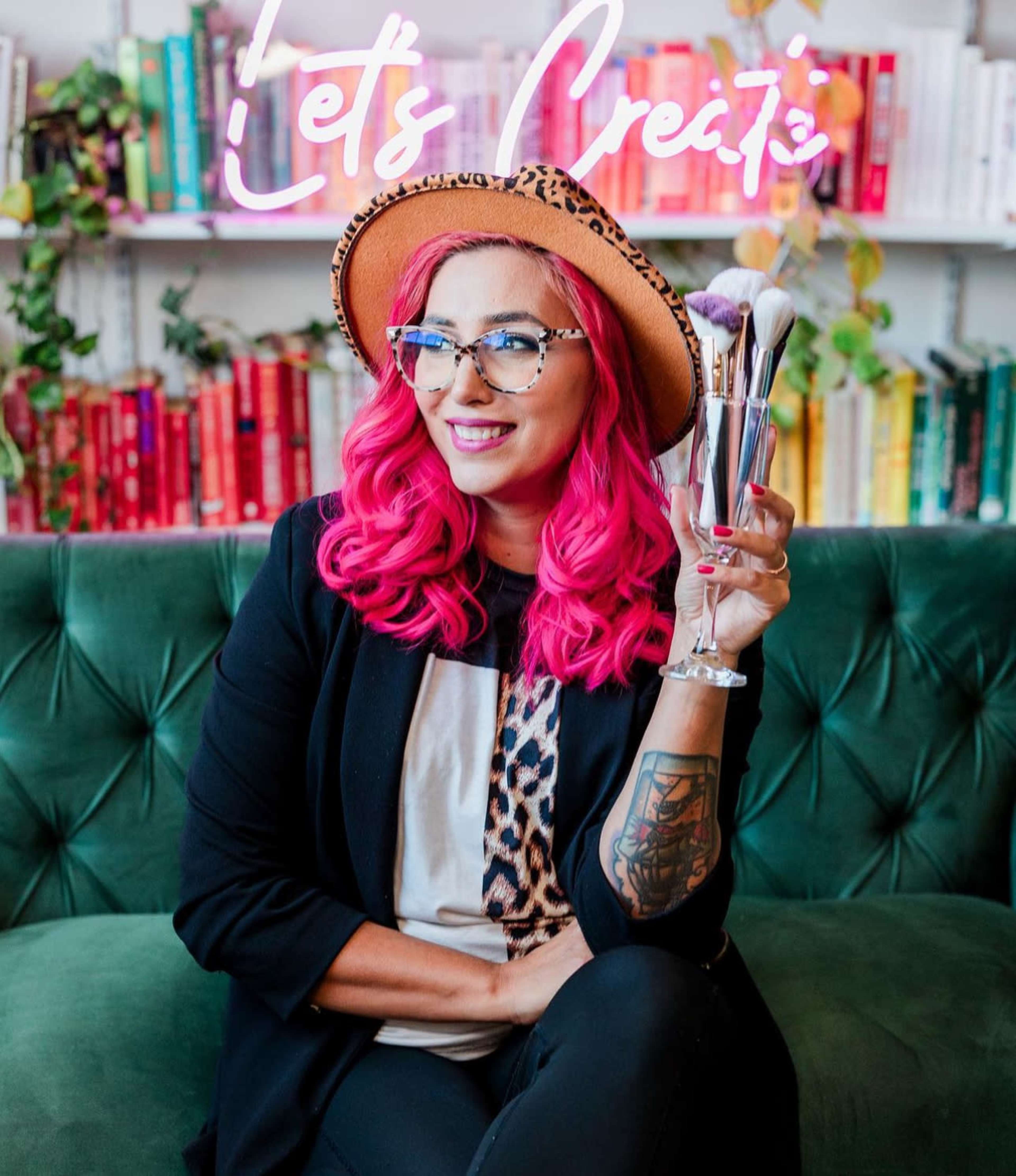 A woman with bright pink hair holds makeup brushes while sitting on a green velvet couch in front of a colorful bookshelf.