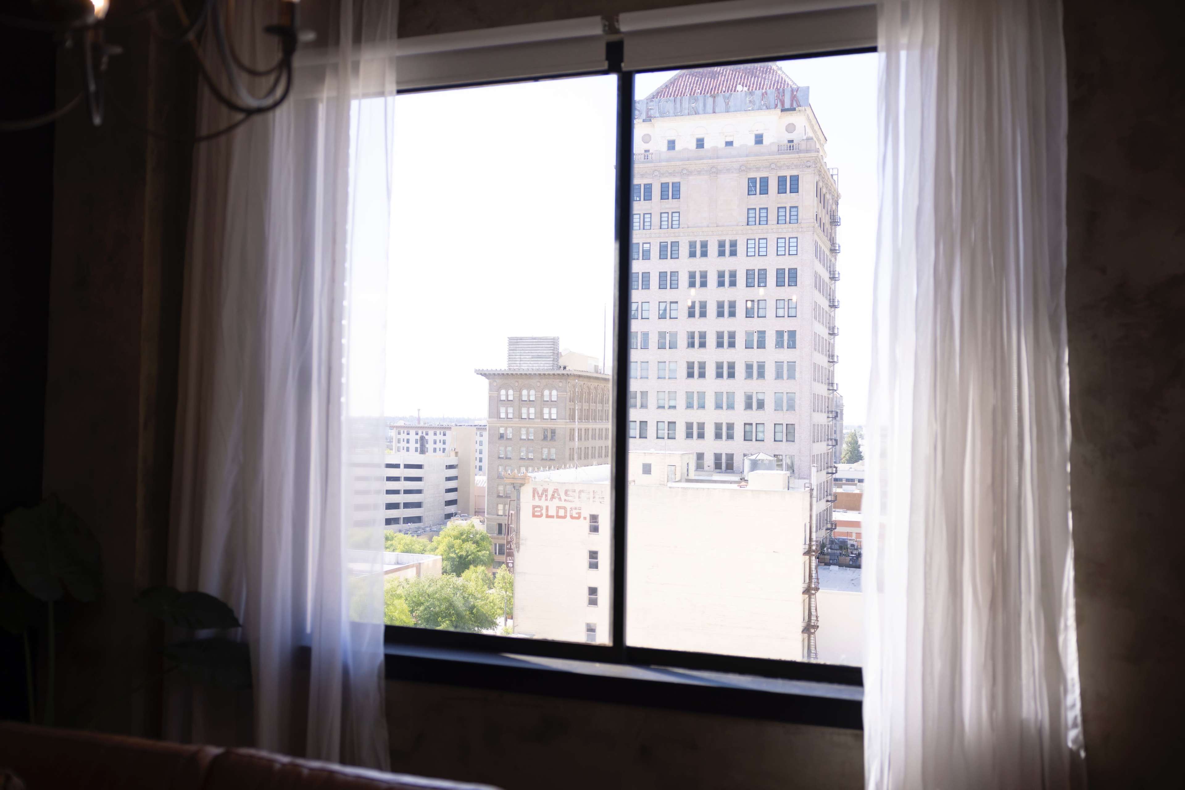 A view from a window with sheer curtains, overlooking a cityscape that includes several buildings.