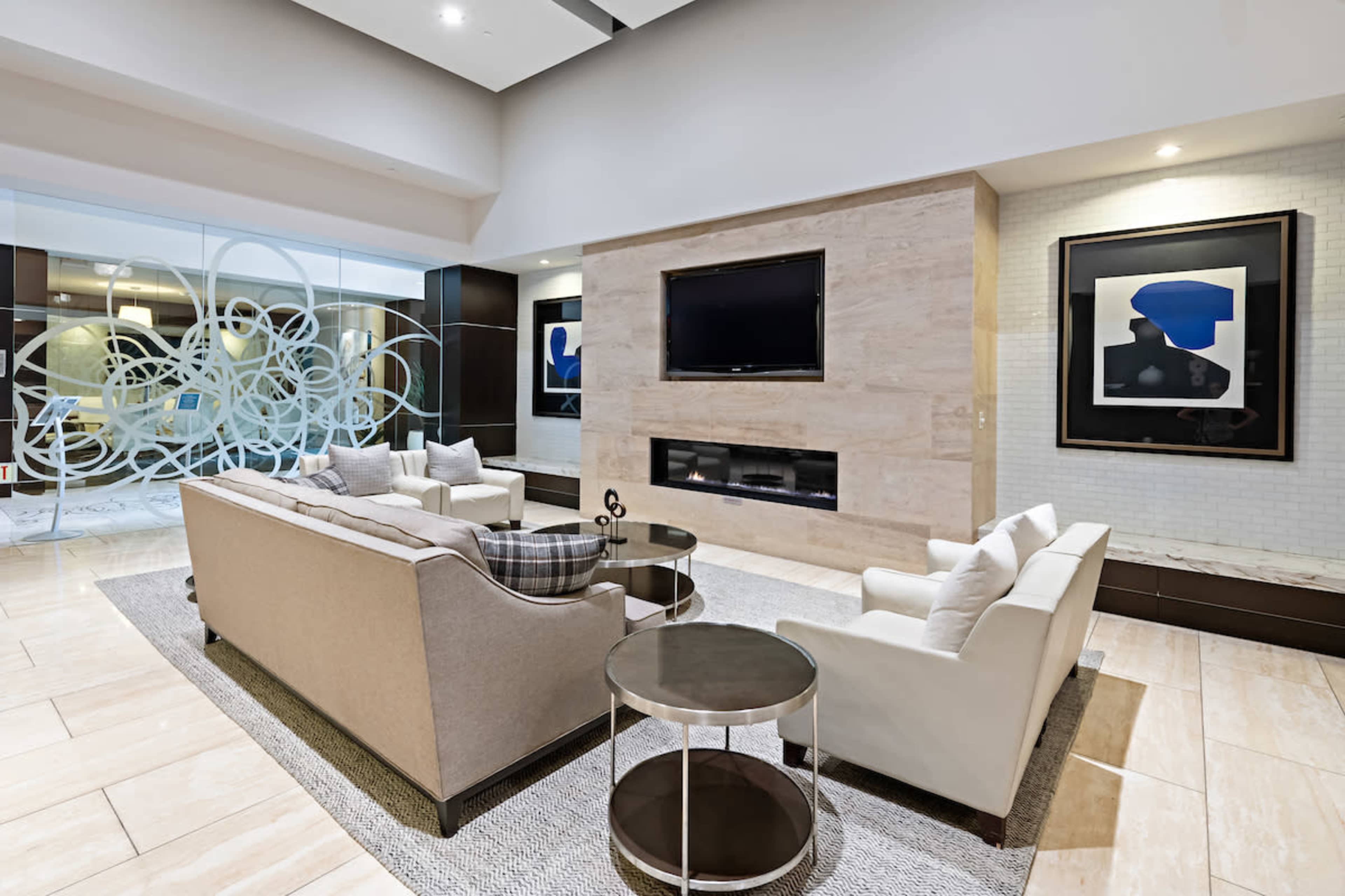 The image shows a modern lobby area featuring two sofas, a round coffee table, a television mounted on a stone wall, and abstract artwork on display.