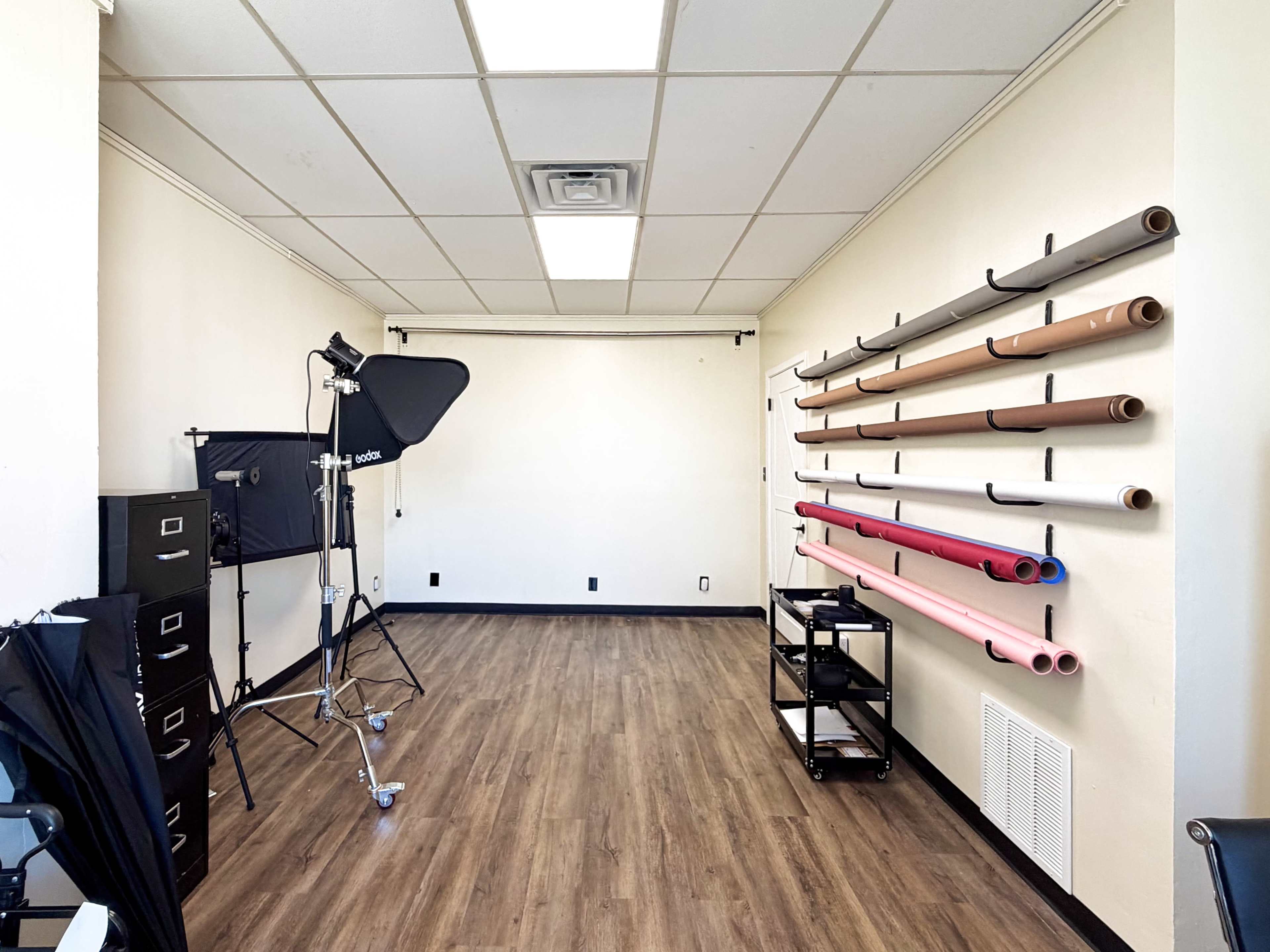 The image shows a spacious studio room with roll paper backdrops mounted on the wall, a lighting setup in the corner, and a mobile cart.
