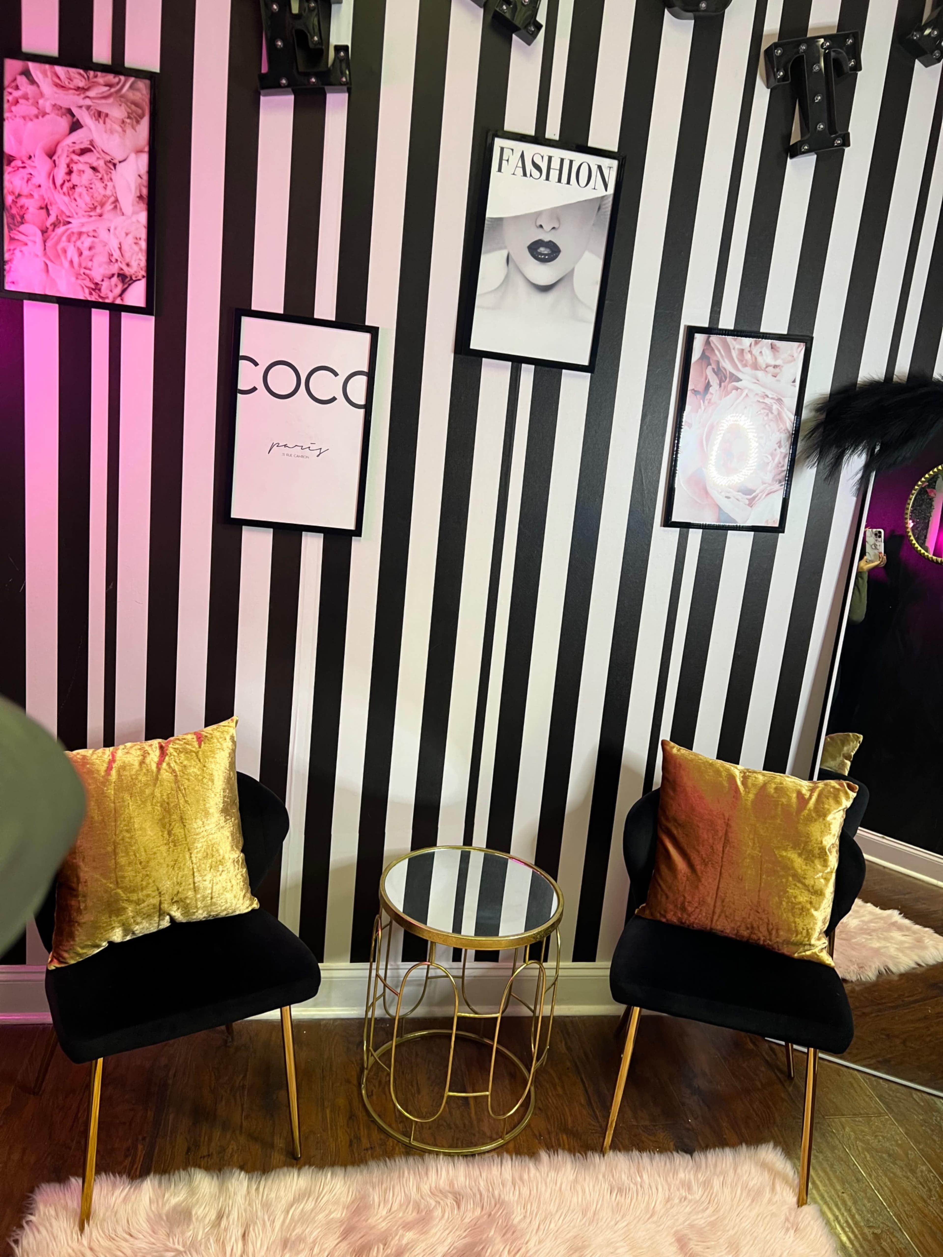The image shows a stylish corner with two black chairs adorned with golden pillows, a round glass-top table, and a striped wall featuring various fashion-themed artwork.