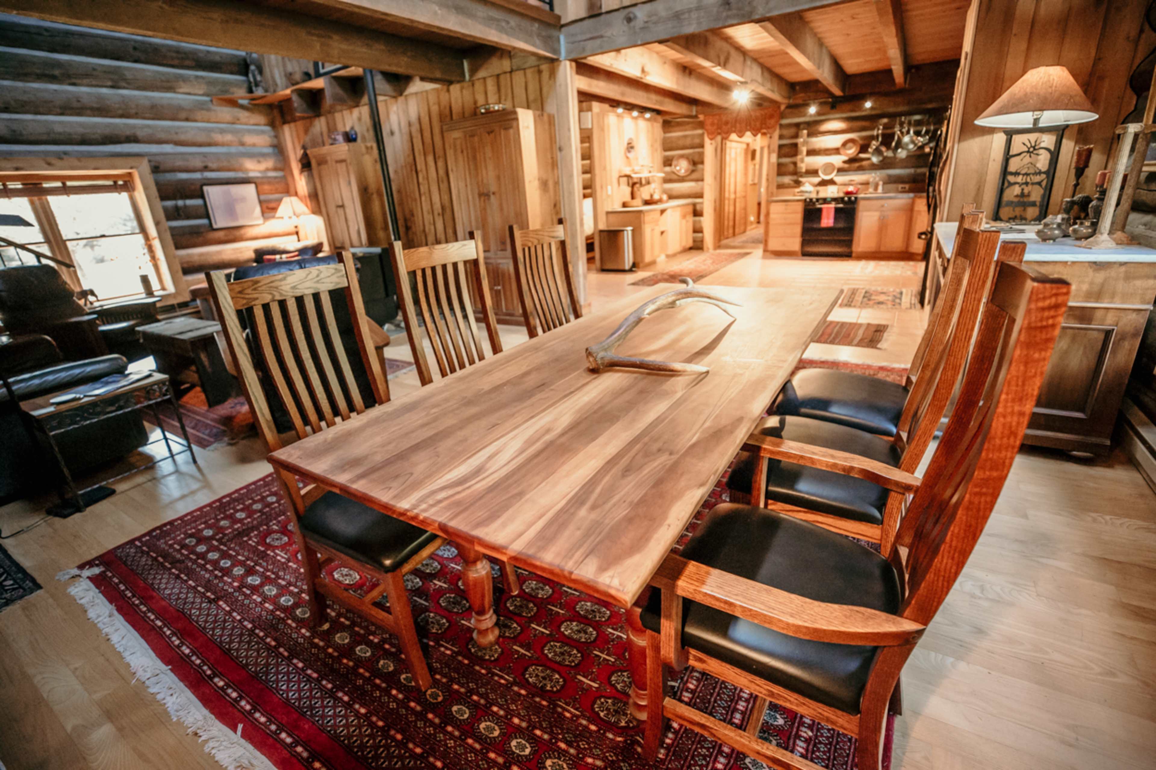 A spacious wooden dining area features a long table surrounded by several chairs, with a traditional rug on the floor and a cozy atmosphere enhanced by wooden beams and light fixtures.