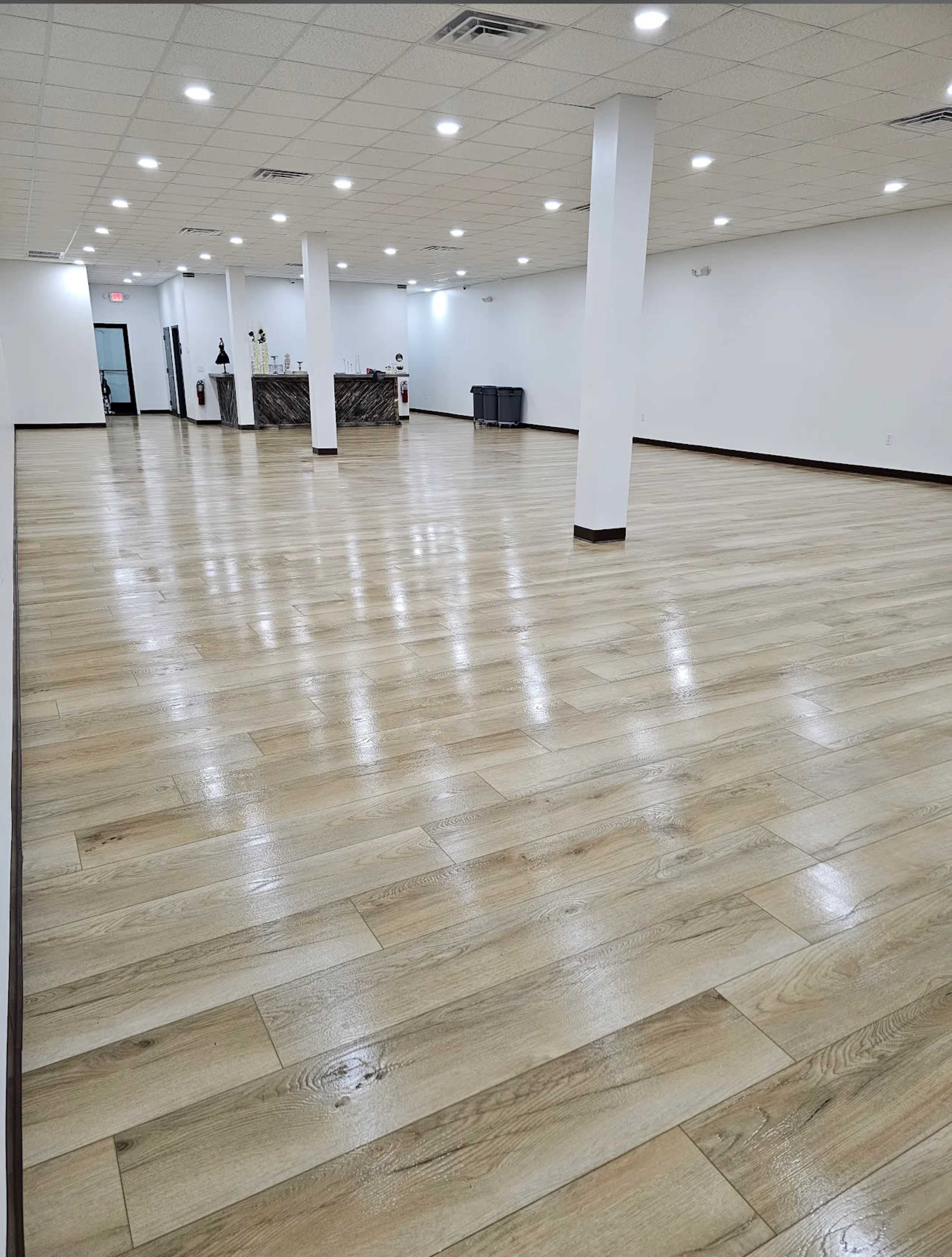 The image shows a spacious, empty room with polished wooden floors, white walls, and a reception area visible in the background.