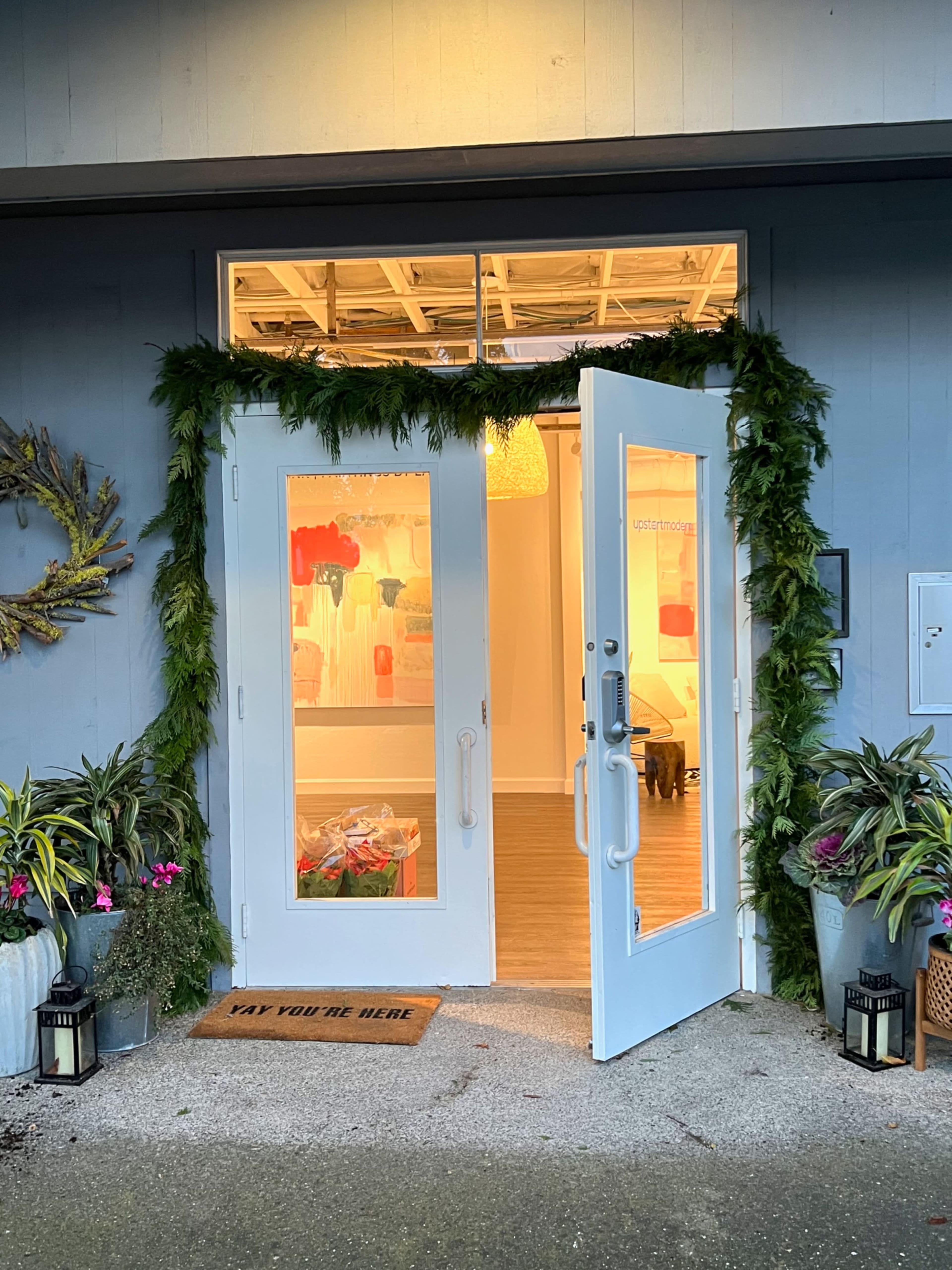 A storefront with double white doors framed by greenery and potted plants, featuring a welcome mat that reads "YAY YOU'RE HERE."