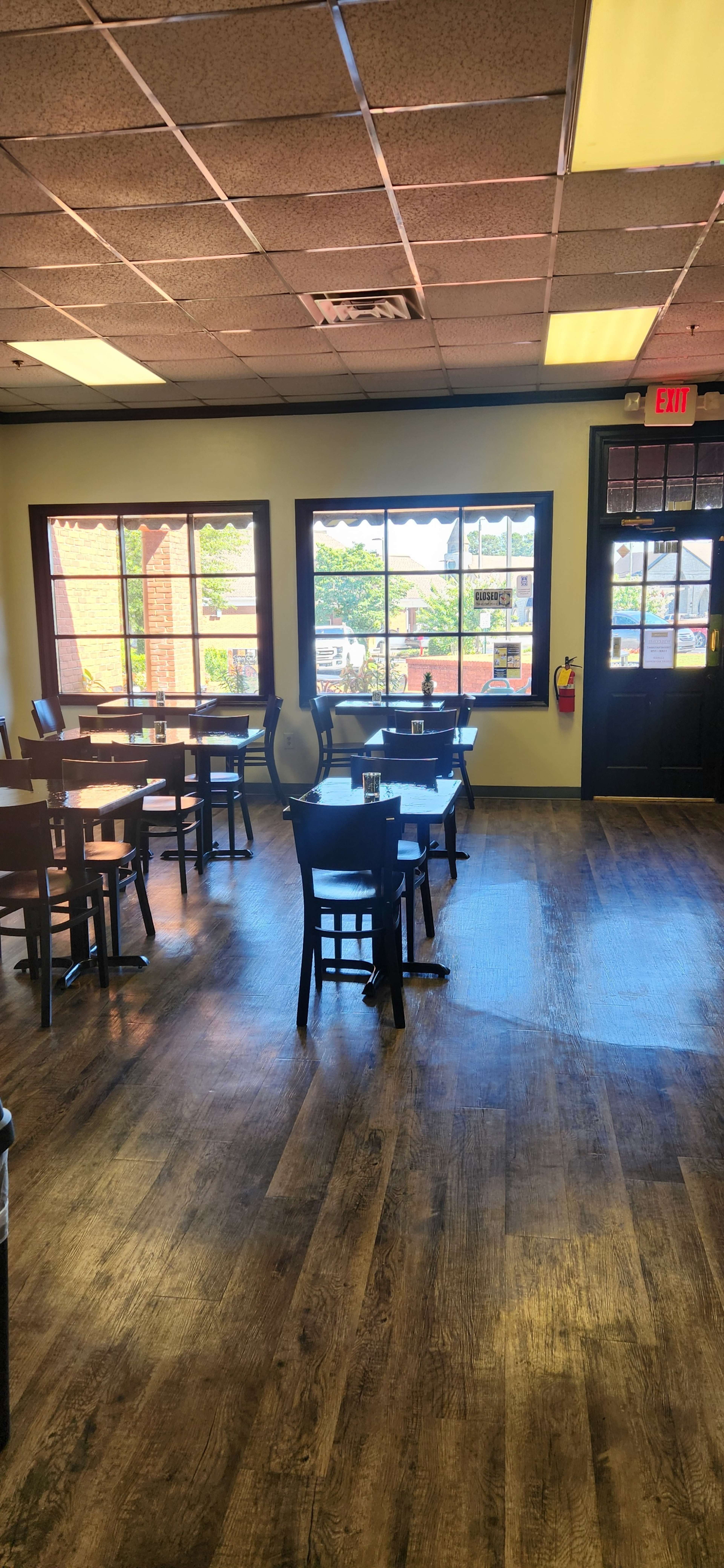 The image shows a partially filled dining room with wooden tables and chairs, large windows illuminating the space, and an exit door on the right.