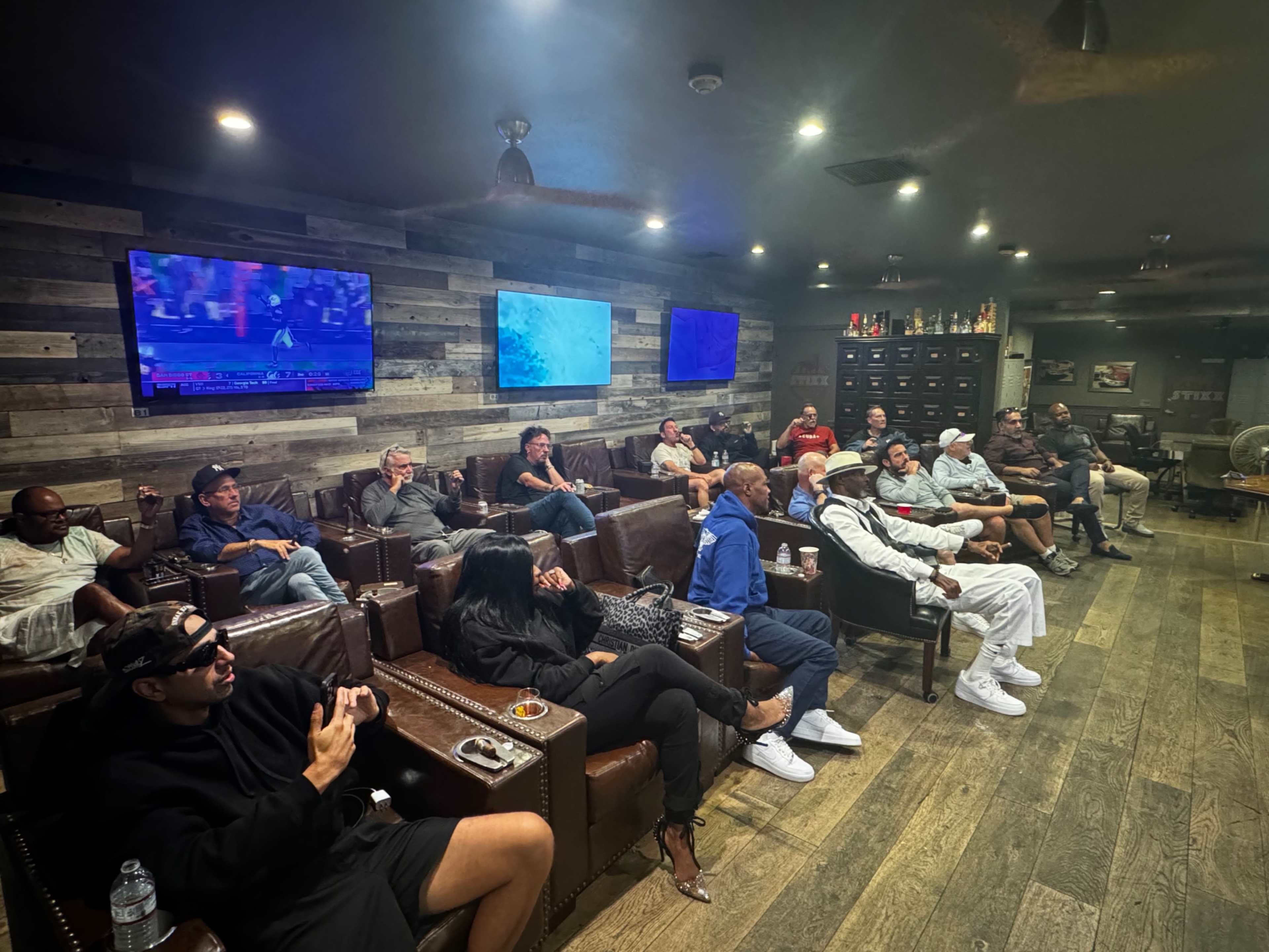A group of people sits in a dimly lit lounge with multiple TVs on the walls, watching a sports event while seated in leather chairs.
