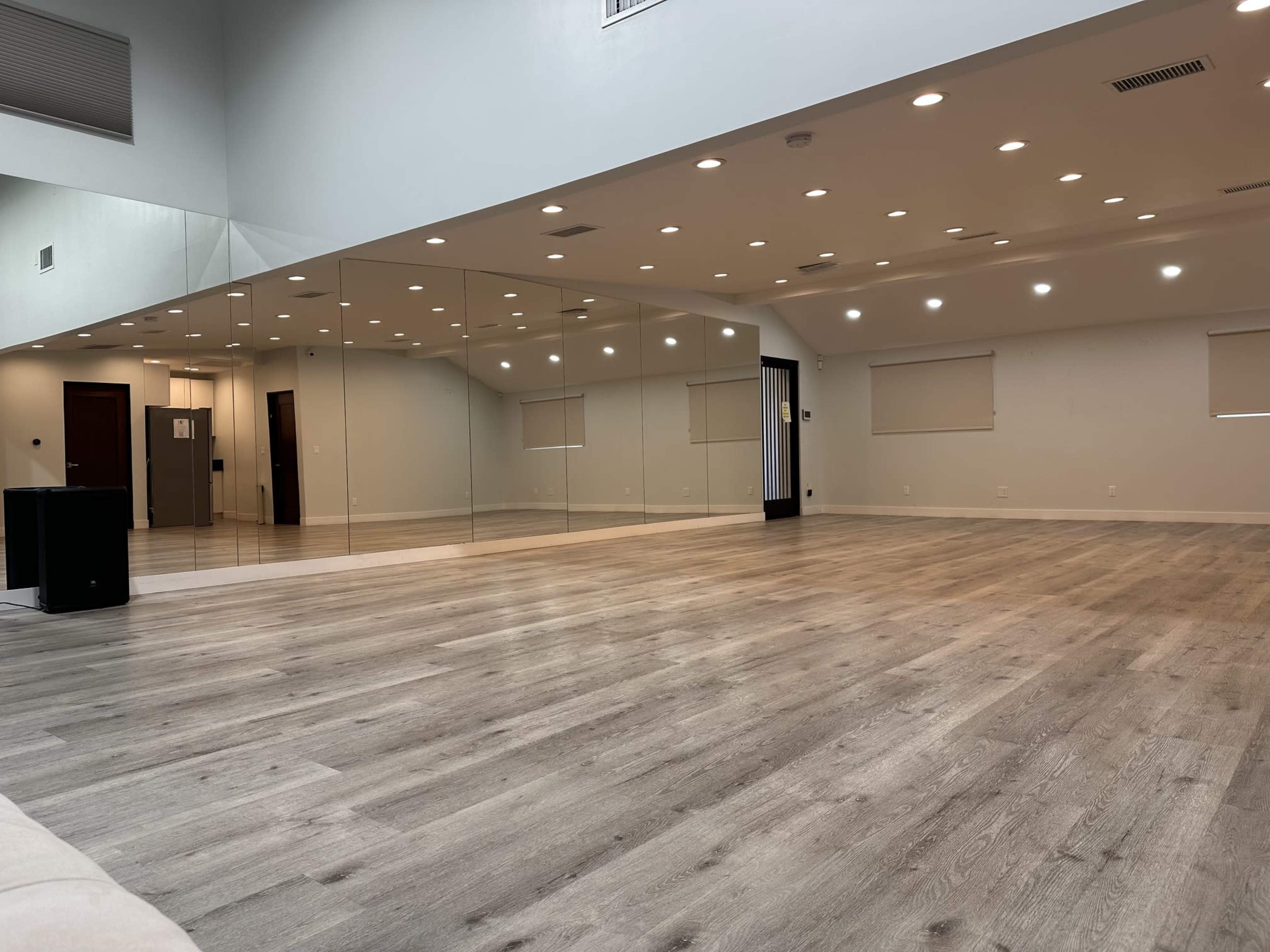 The image shows a spacious room with wooden flooring, large mirrors on one wall, and bright ceiling lights.