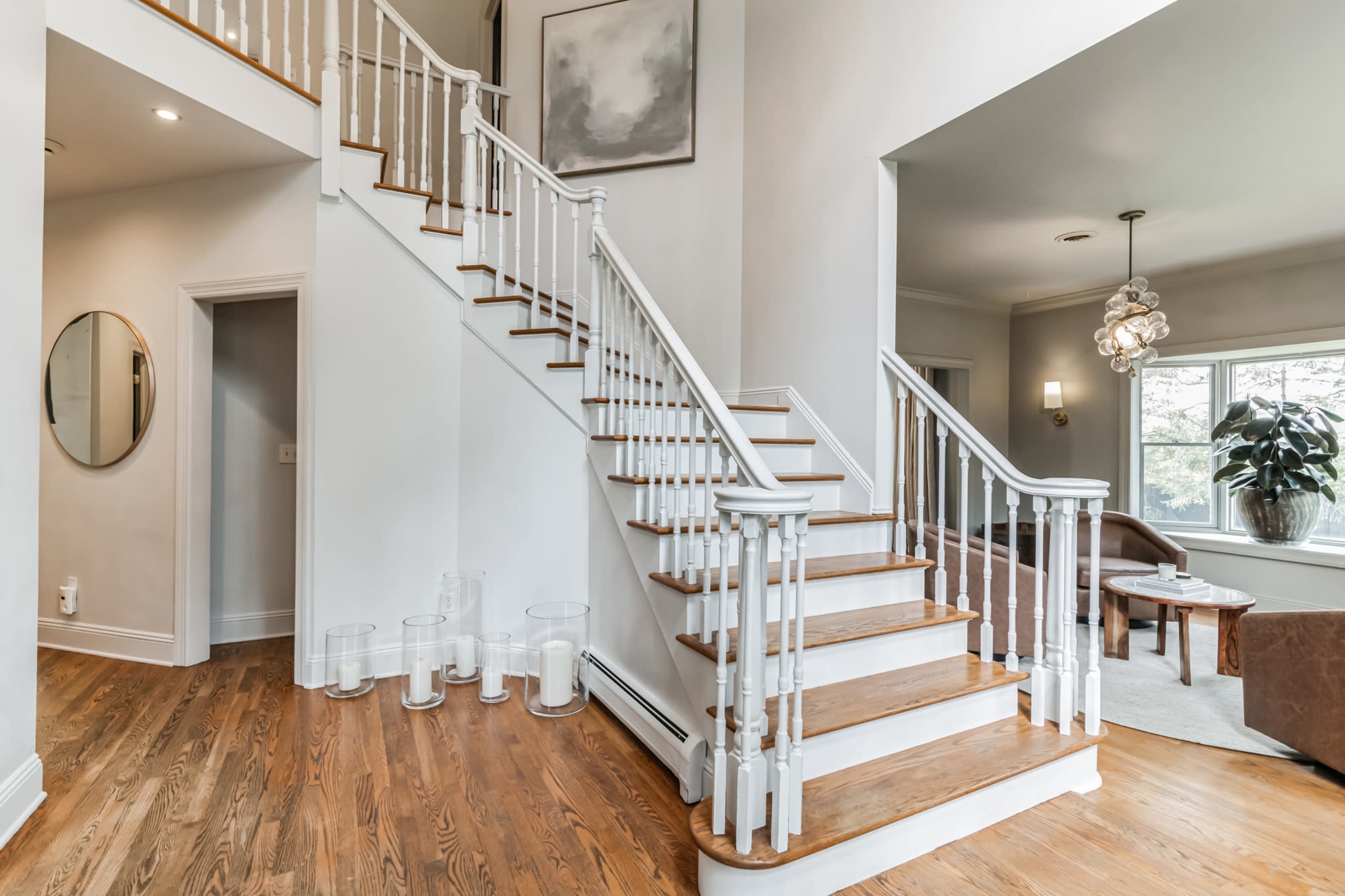 A well-lit foyer features a staircase with white railings and wooden steps, flanked by a circular mirror and decorative candles on the floor.