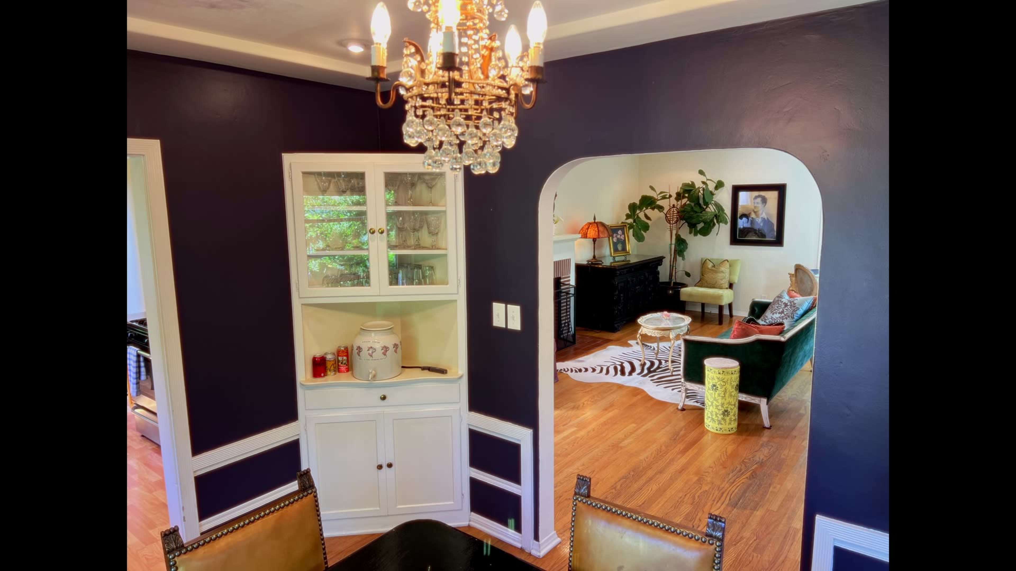 The image shows a dining area with a chandelier and a white cabinet, leading to a living room decorated with a zebra-striped rug and green furniture.