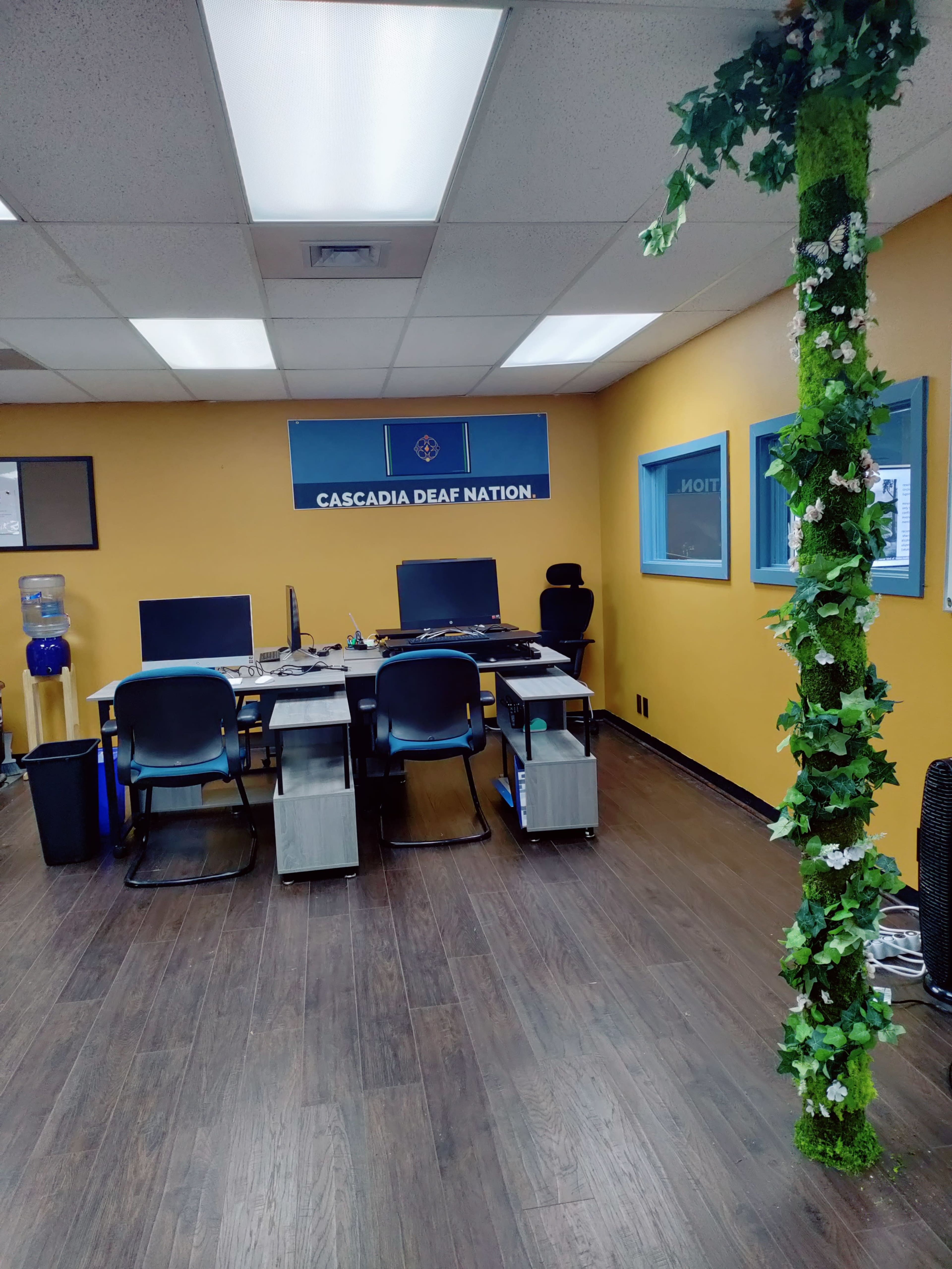 An office space with a yellow wall, a "Cascadia Deaf Nation" sign, two desks with computers, and a green plant-covered column.