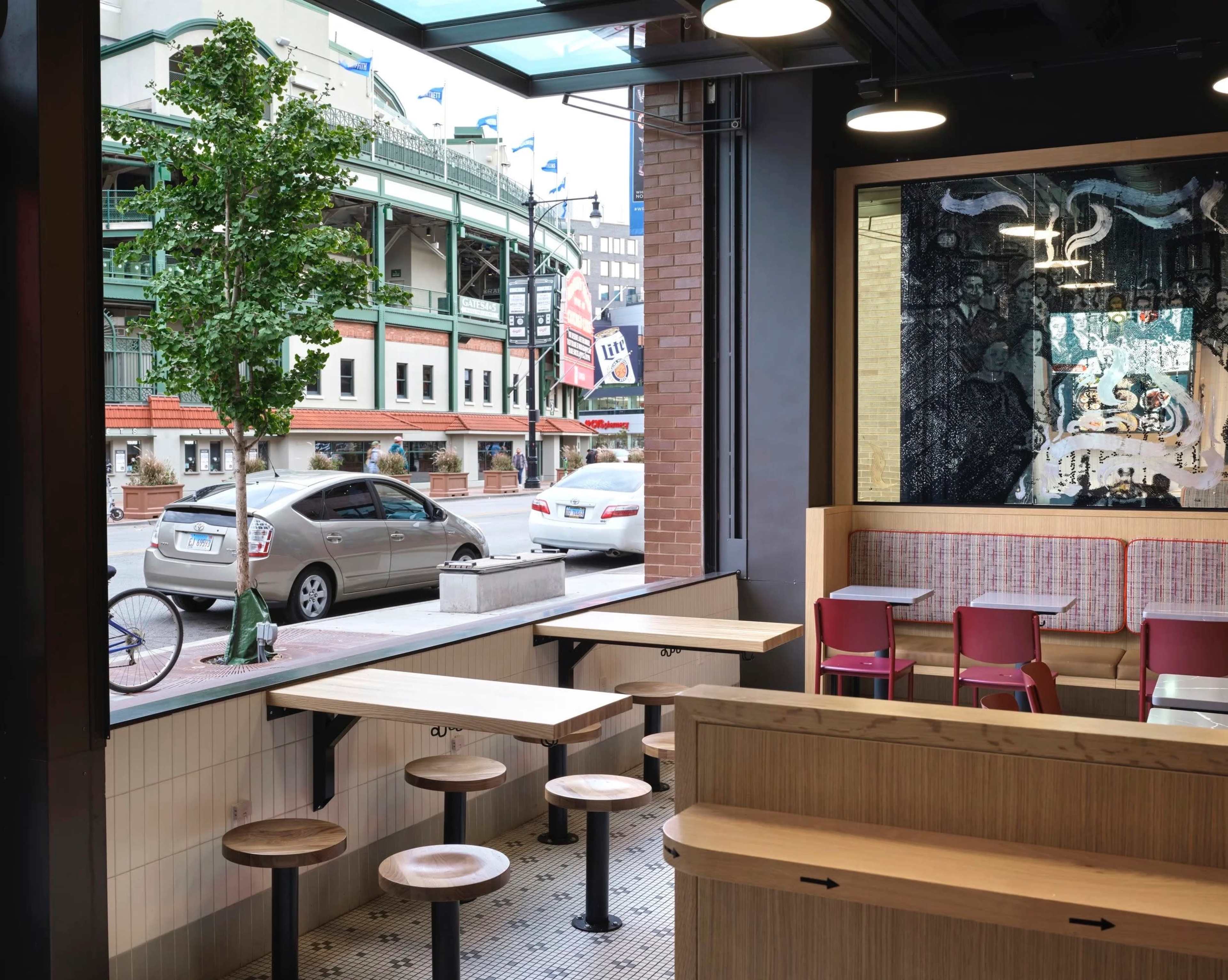 The image shows the interior of a modern restaurant with wooden tables and chairs, large windows overlooking a street with a building and parked cars in the background.
