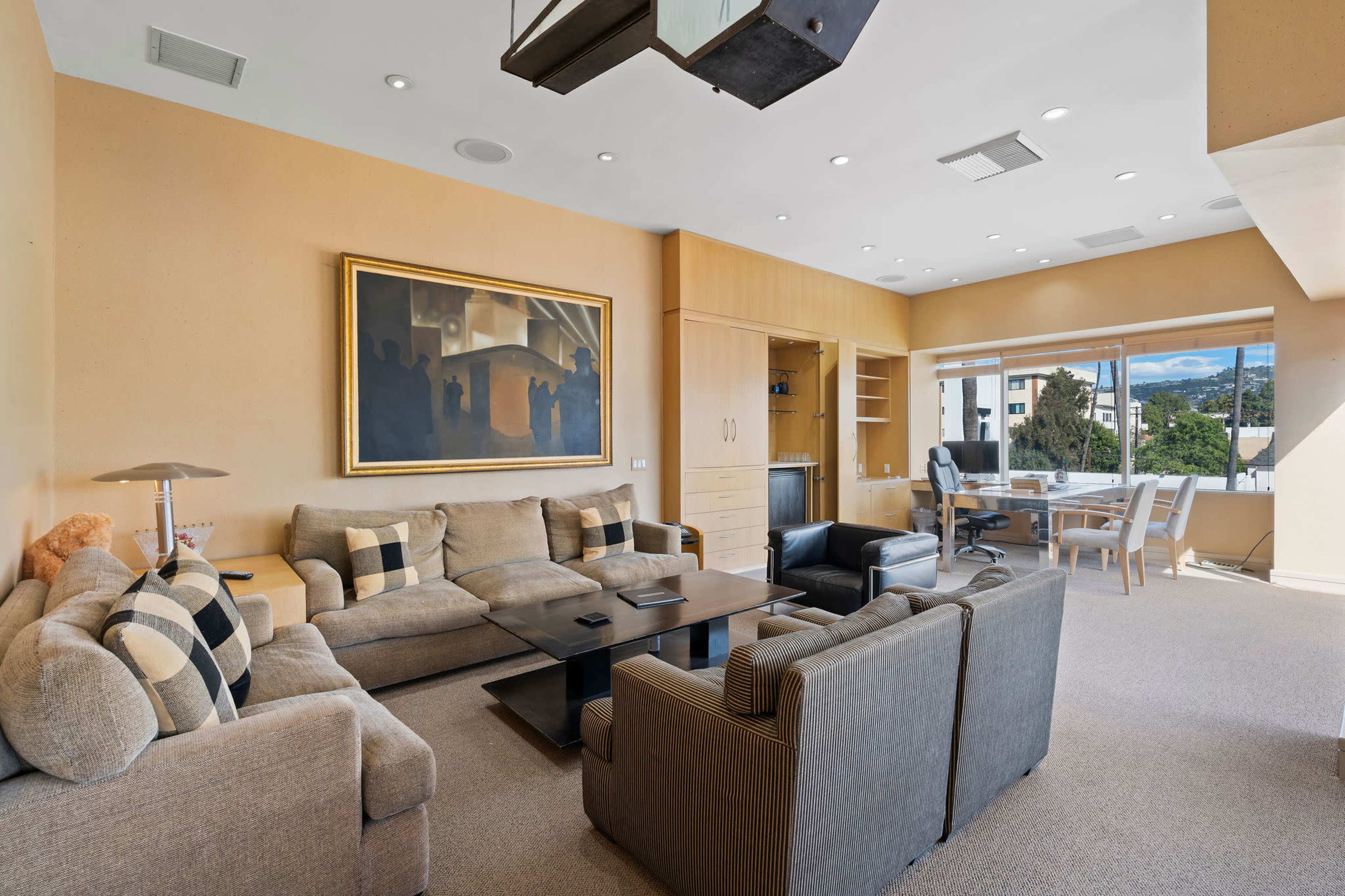 The image shows a modern living room with a large sectional sofa and an adjoining workspace featuring a desk and chairs, illuminated by natural light from large windows.