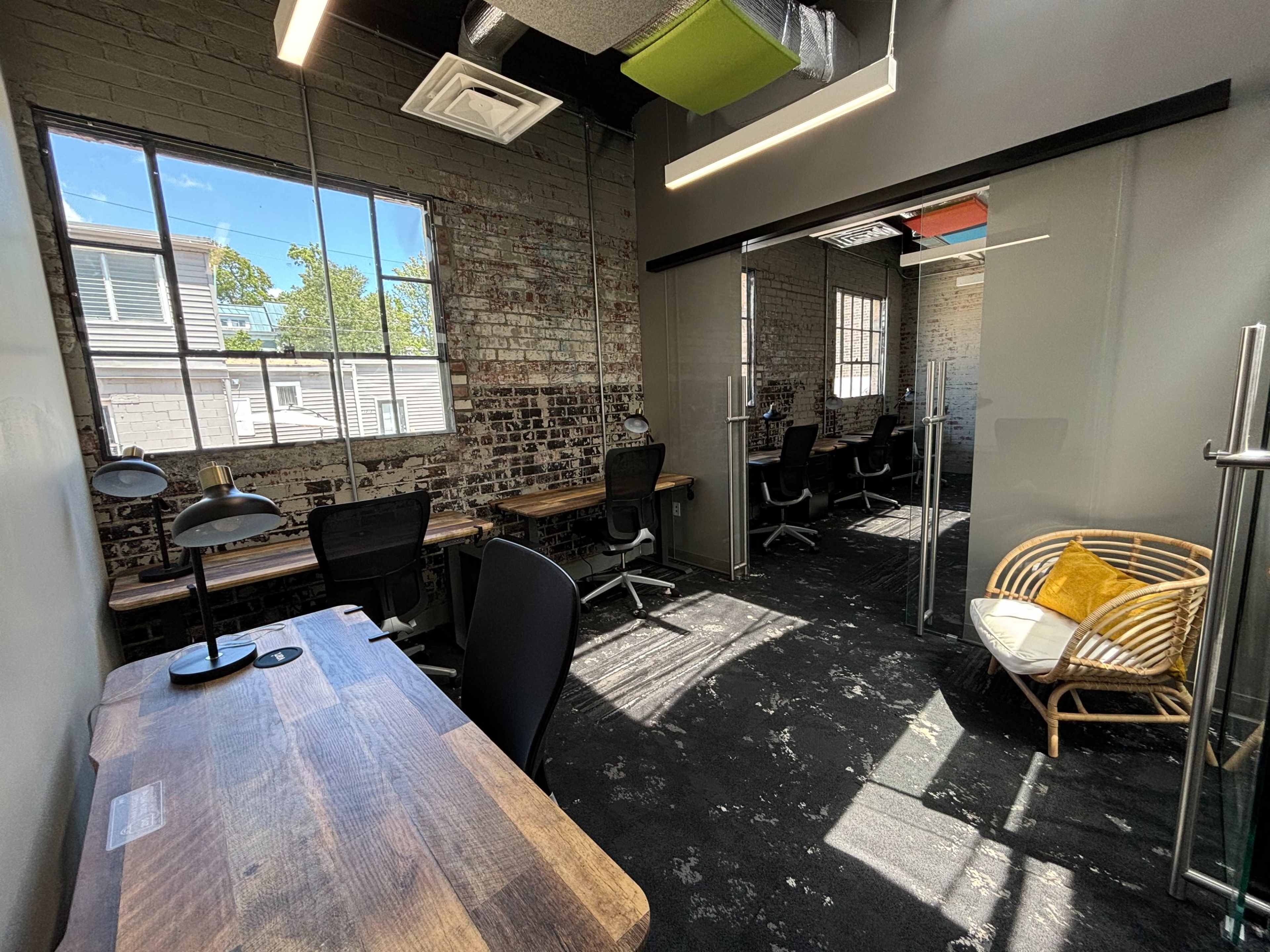 A modern office space with wooden desks, black chairs, and large windows allowing natural light to enter, featuring a cozy seating area with a rattan chair.
