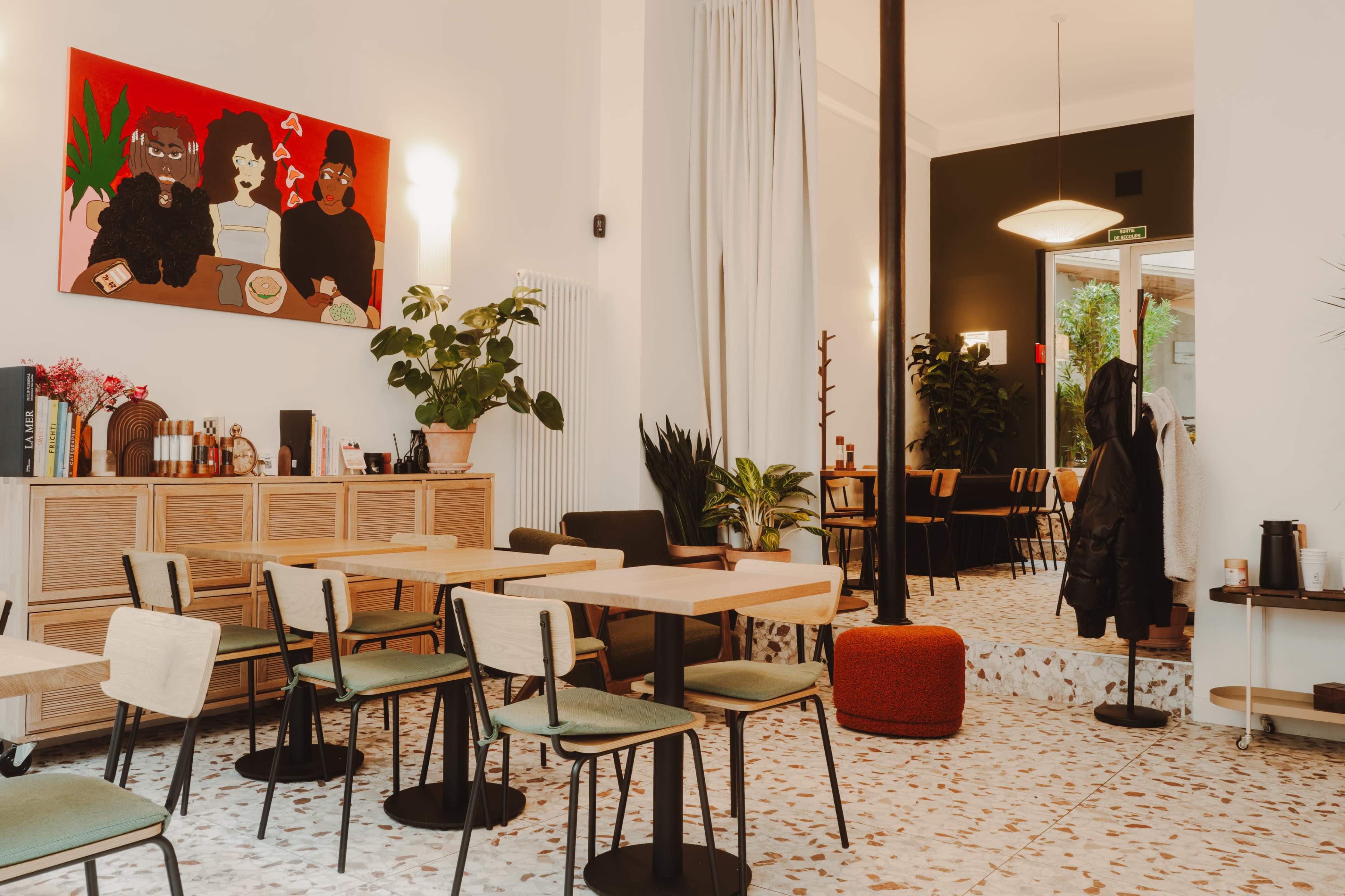 A modern interior of a cafe features wooden tables and chairs, a large colorful artwork on the wall, and indoor plants.