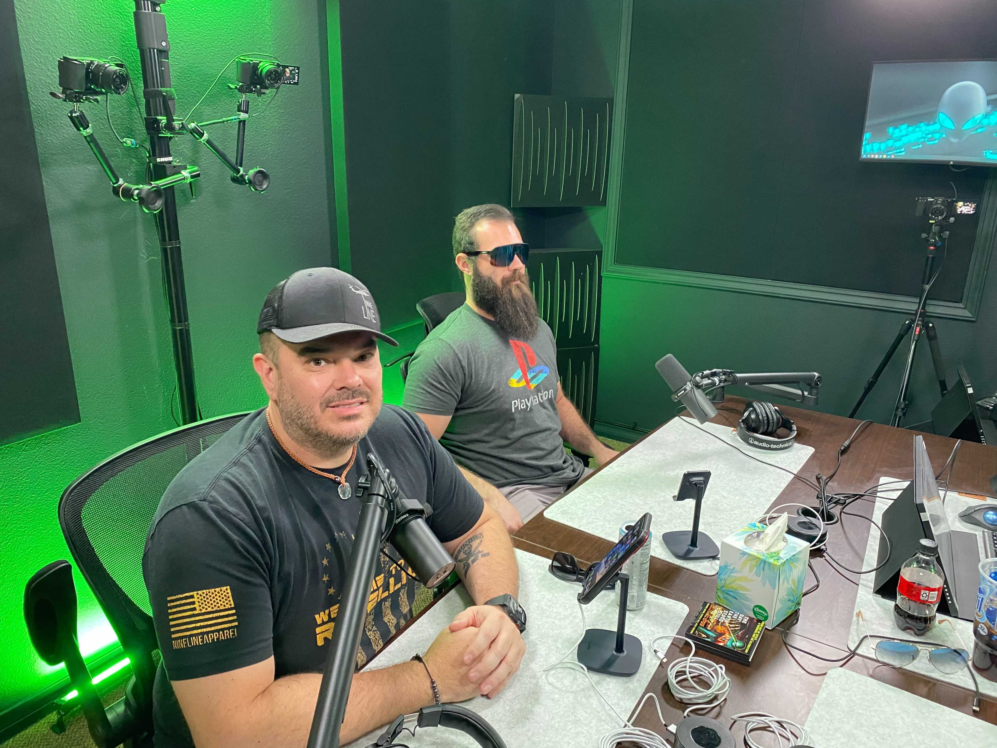Two men are seated at a table in a studio setting, equipped with microphones and recording equipment, with green lighting in the background.