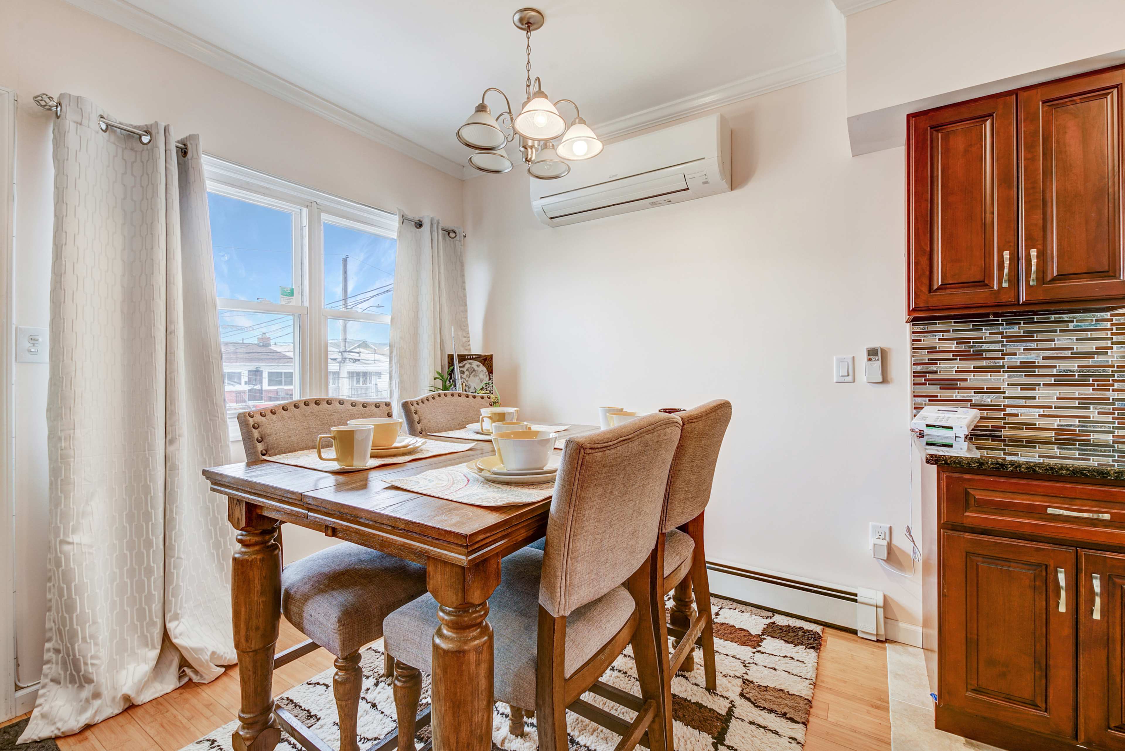 A dining area features a wooden table set with dishware and four upholstered chairs, alongside a window with curtains and an air conditioning unit on the wall.