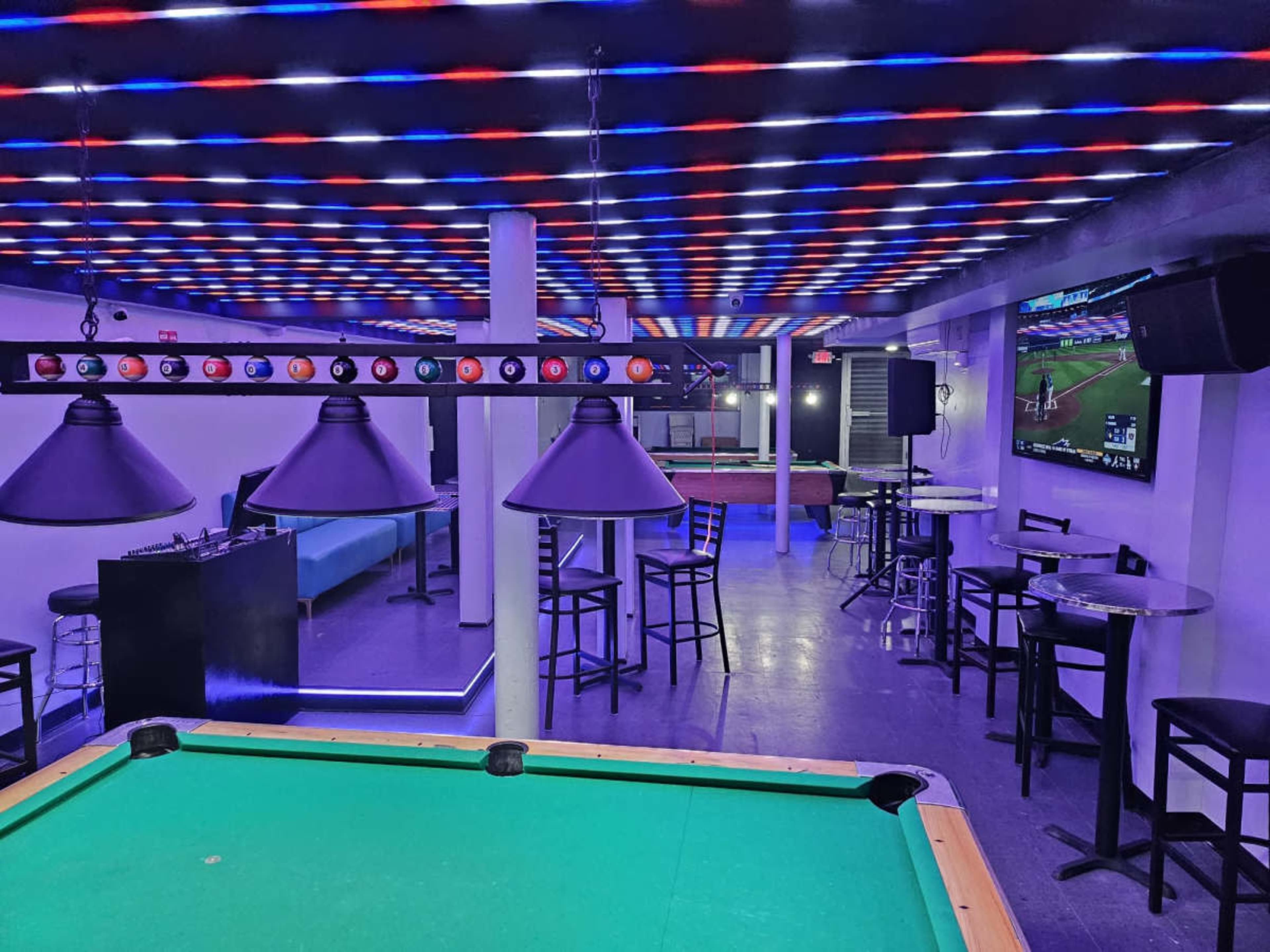 The image shows a modern lounge area featuring a green billiards table, several dark tables and chairs, and colorful ceiling lights.