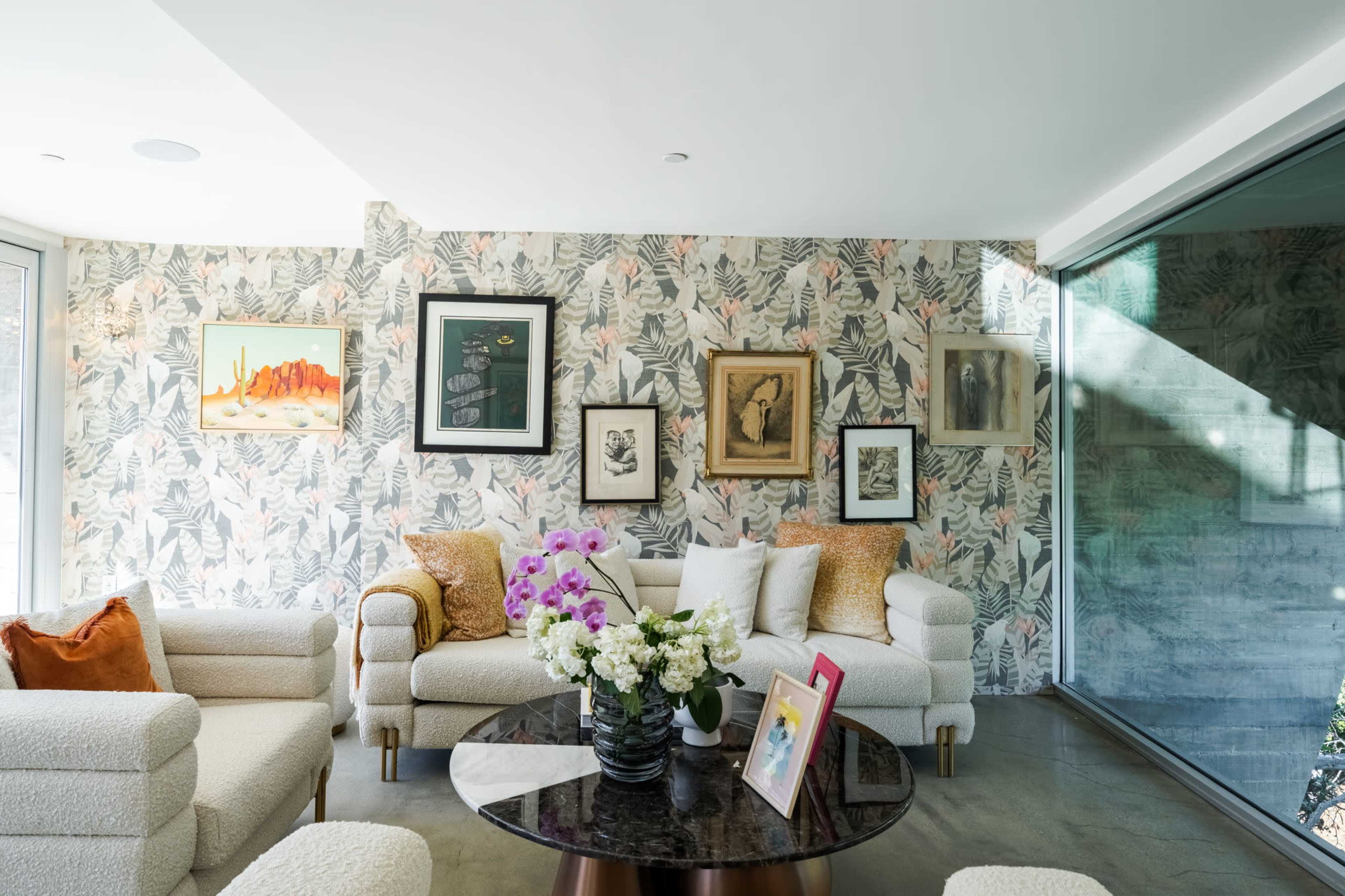 The image shows a modern living room with light-colored sofas, a dark round coffee table, floral wallpaper, and framed artwork on the walls.