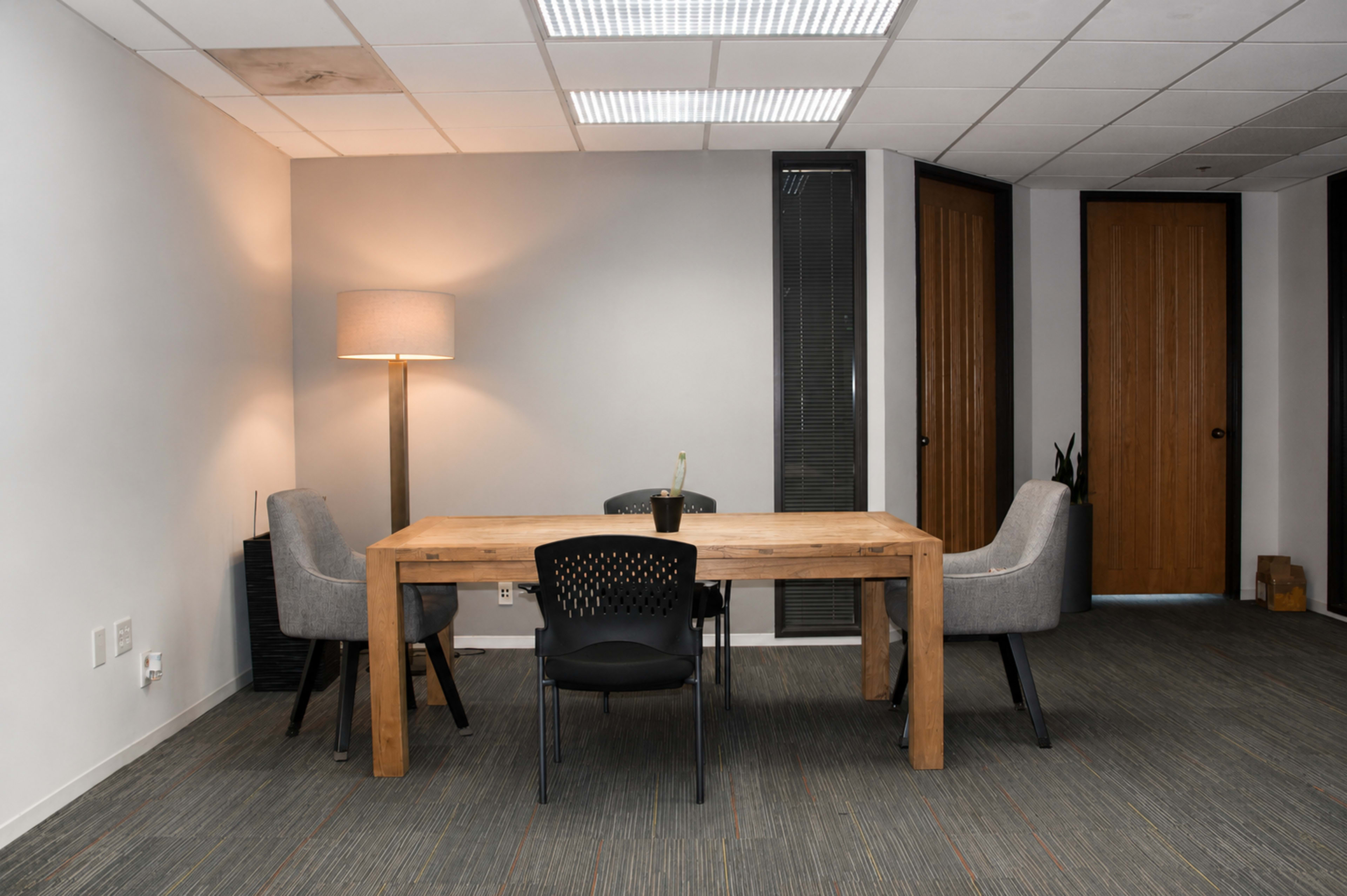 The image shows a minimalist office meeting space featuring a wooden table surrounded by four chairs, with a floor lamp and two closed doors in the background.