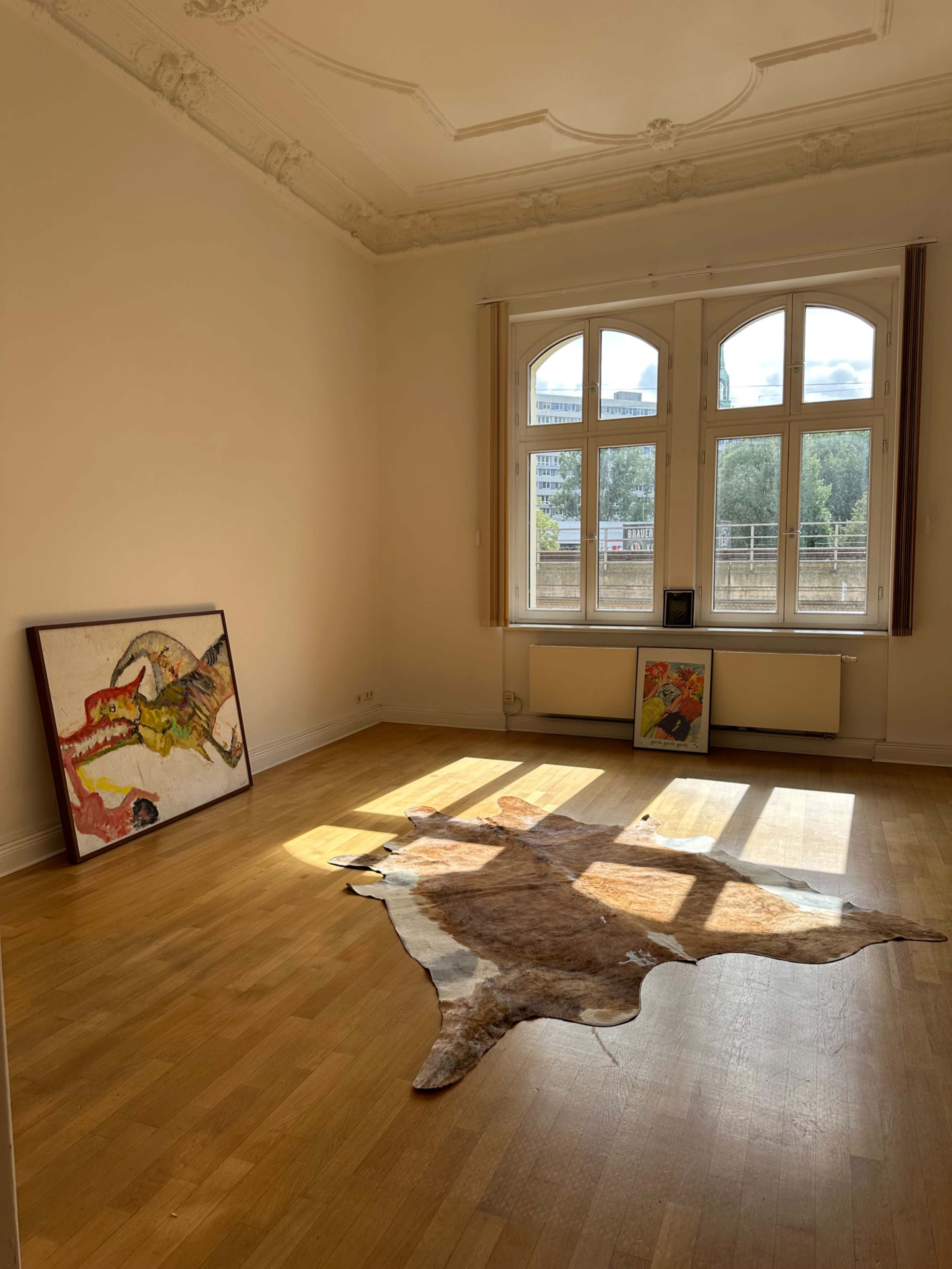 The image shows a bright, empty room with large windows, wooden floors, and two framed paintings leaning against the walls, along with a cowhide rug on the floor.
