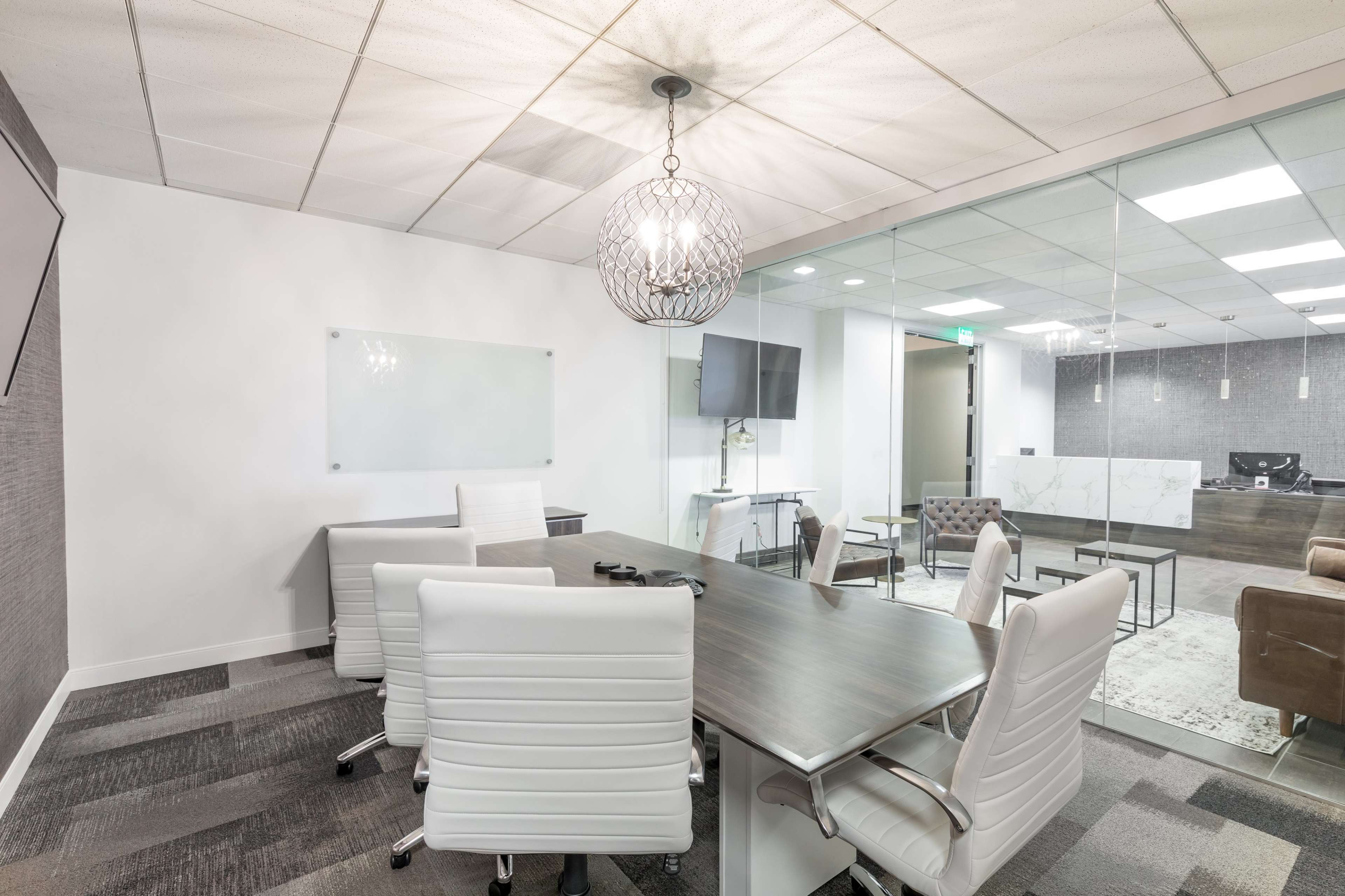 A modern conference room features a large table surrounded by white chairs, with a glass wall leading to an adjoining office space equipped with a desk and television.