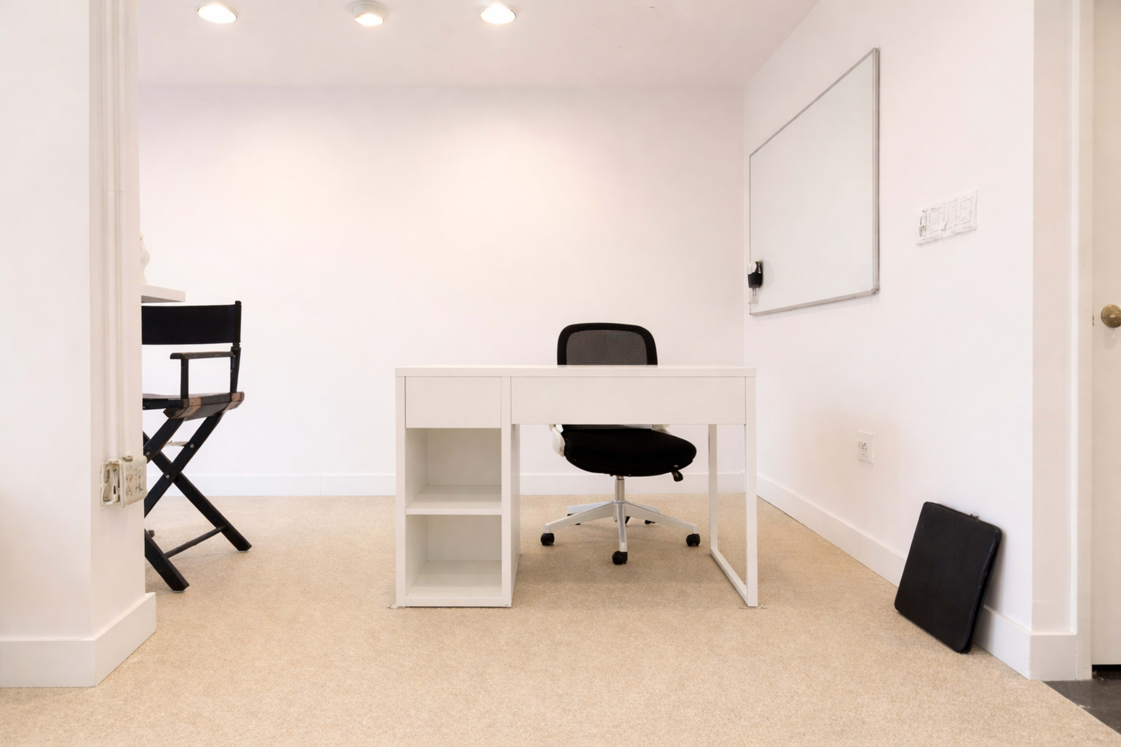 The image shows a minimalist office space featuring a white desk with a black office chair and a nearby empty chair.