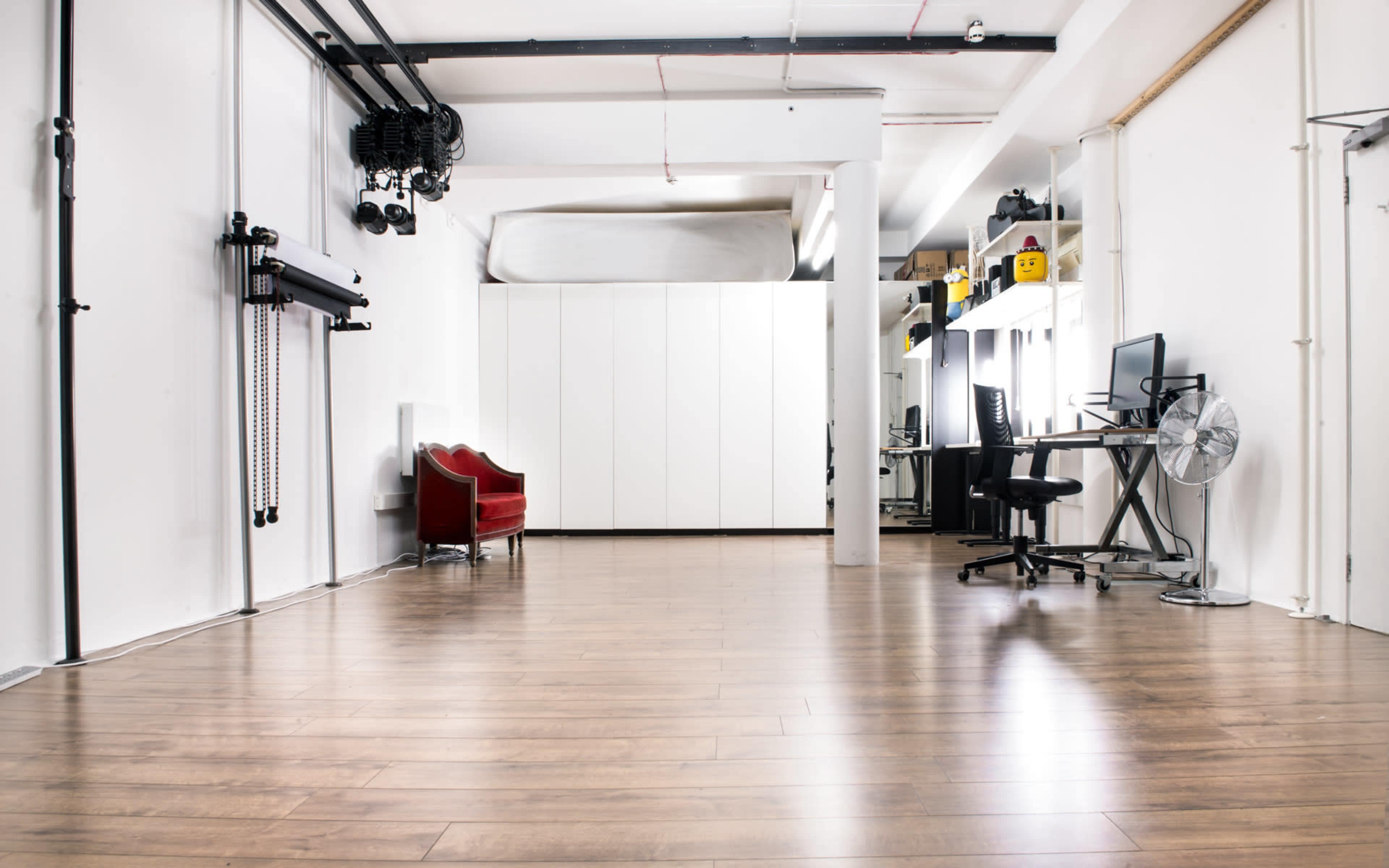 A spacious, minimalist interior with wooden flooring, featuring a red armchair, a white storage unit, and workstations with chairs and a fan.