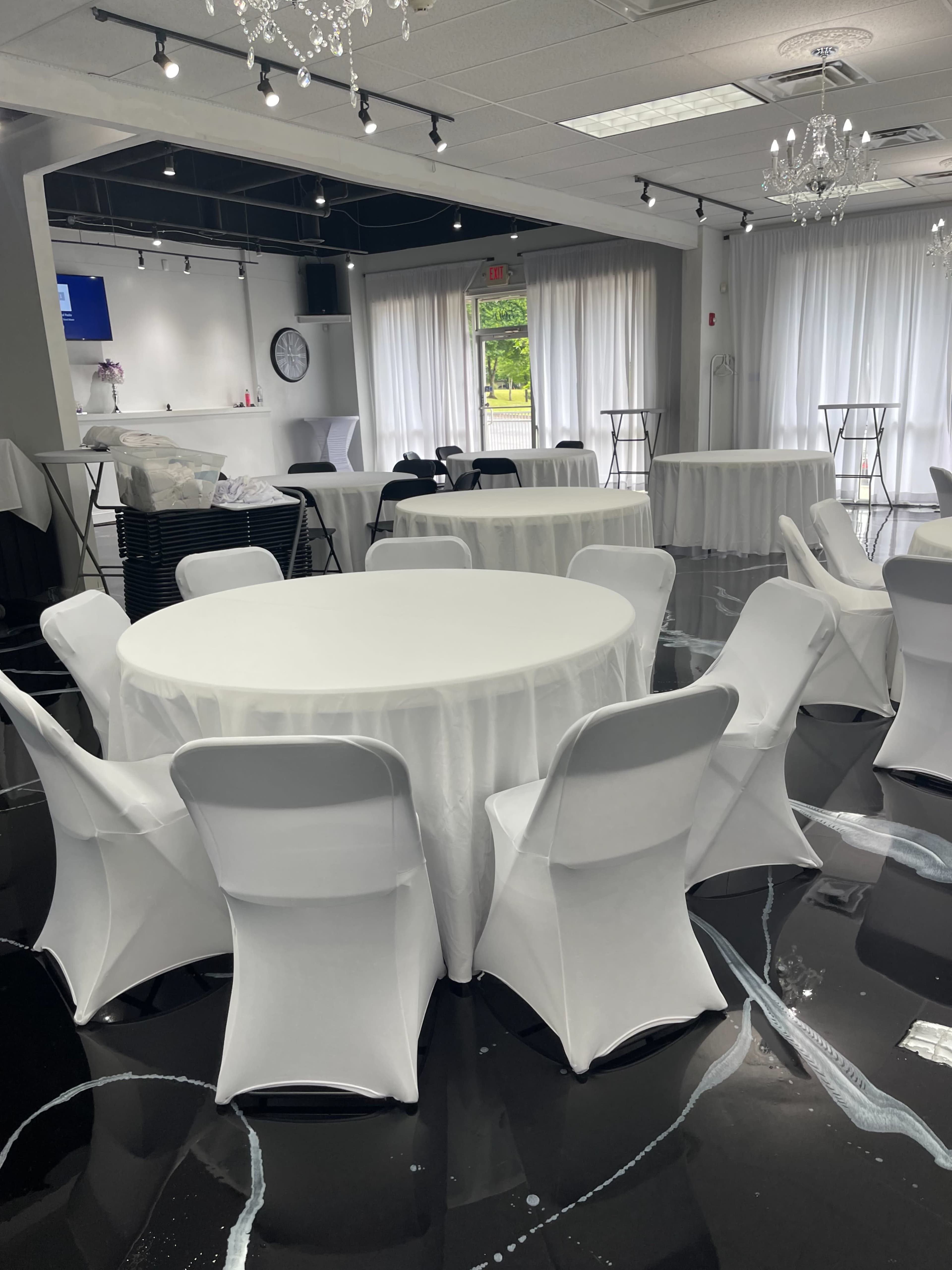 The image shows a spacious event venue with several round tables covered in white tablecloths and white chairs arranged around them, alongside a shiny black floor.