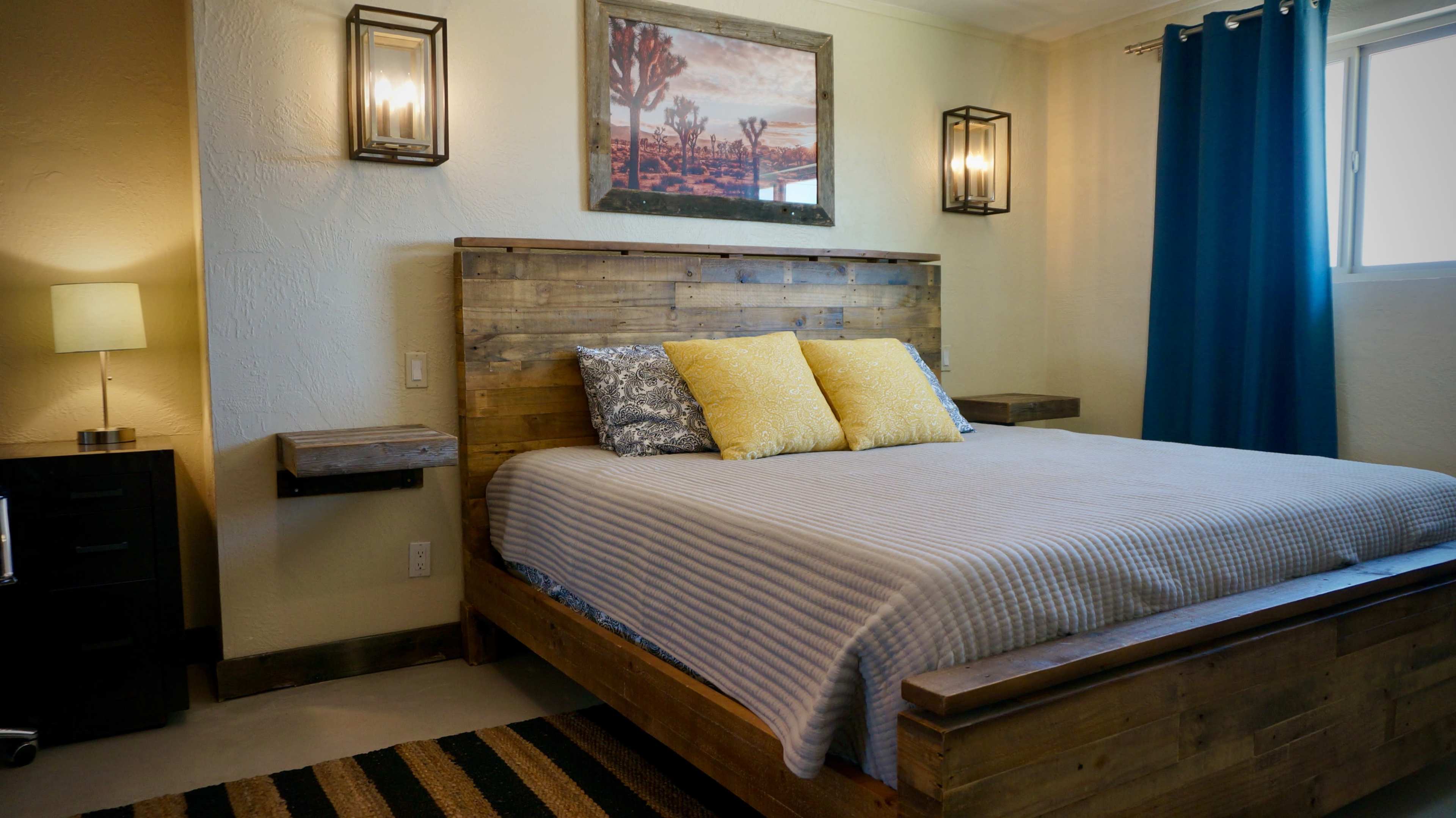 A bedroom featuring a wooden bed frame with yellow pillows, framed art on the wall, and blue curtains.