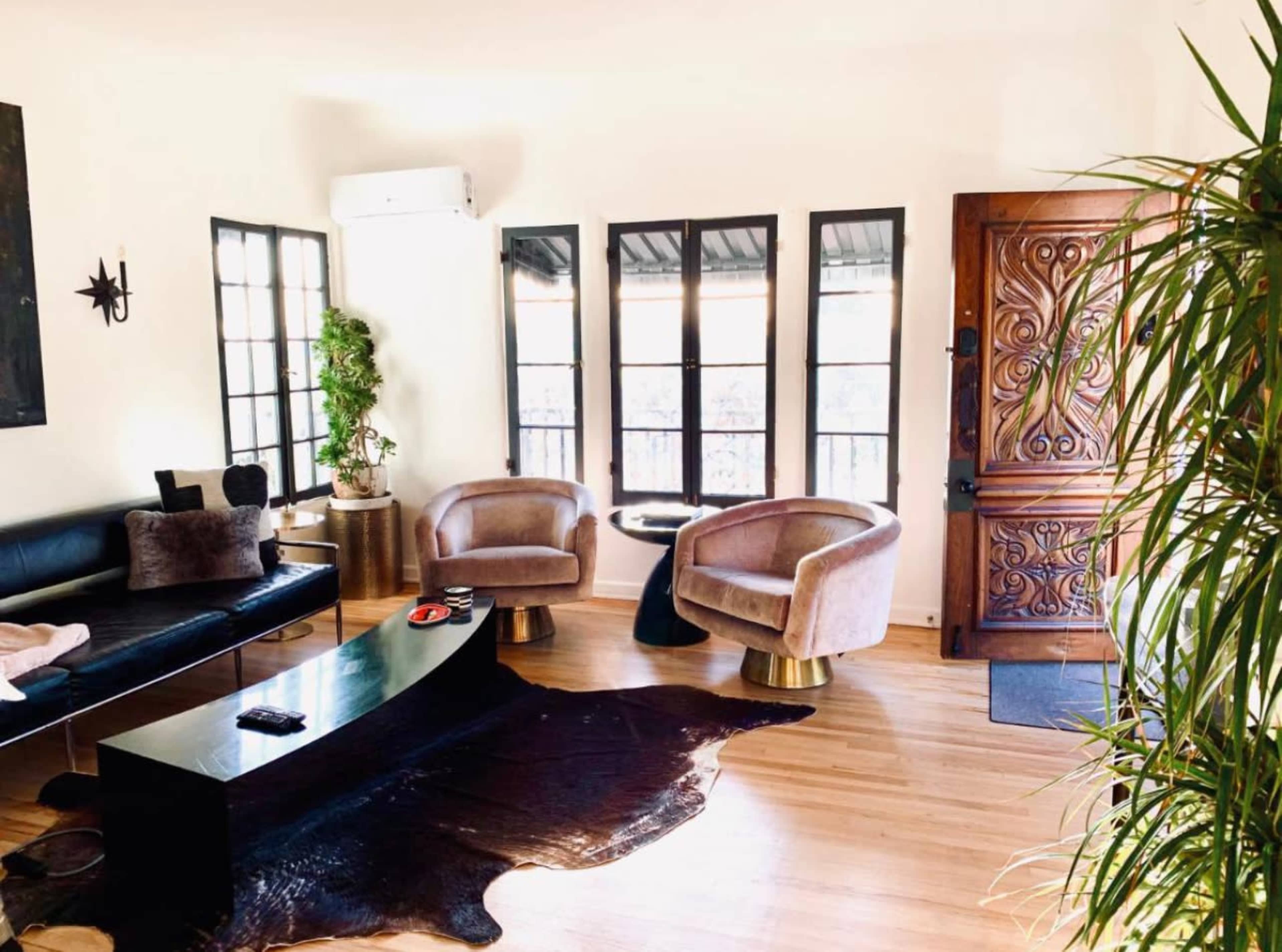 The image shows a modern living room with a black leather sofa, two light-colored armchairs, a sleek coffee table, and large windows allowing natural light into the space.