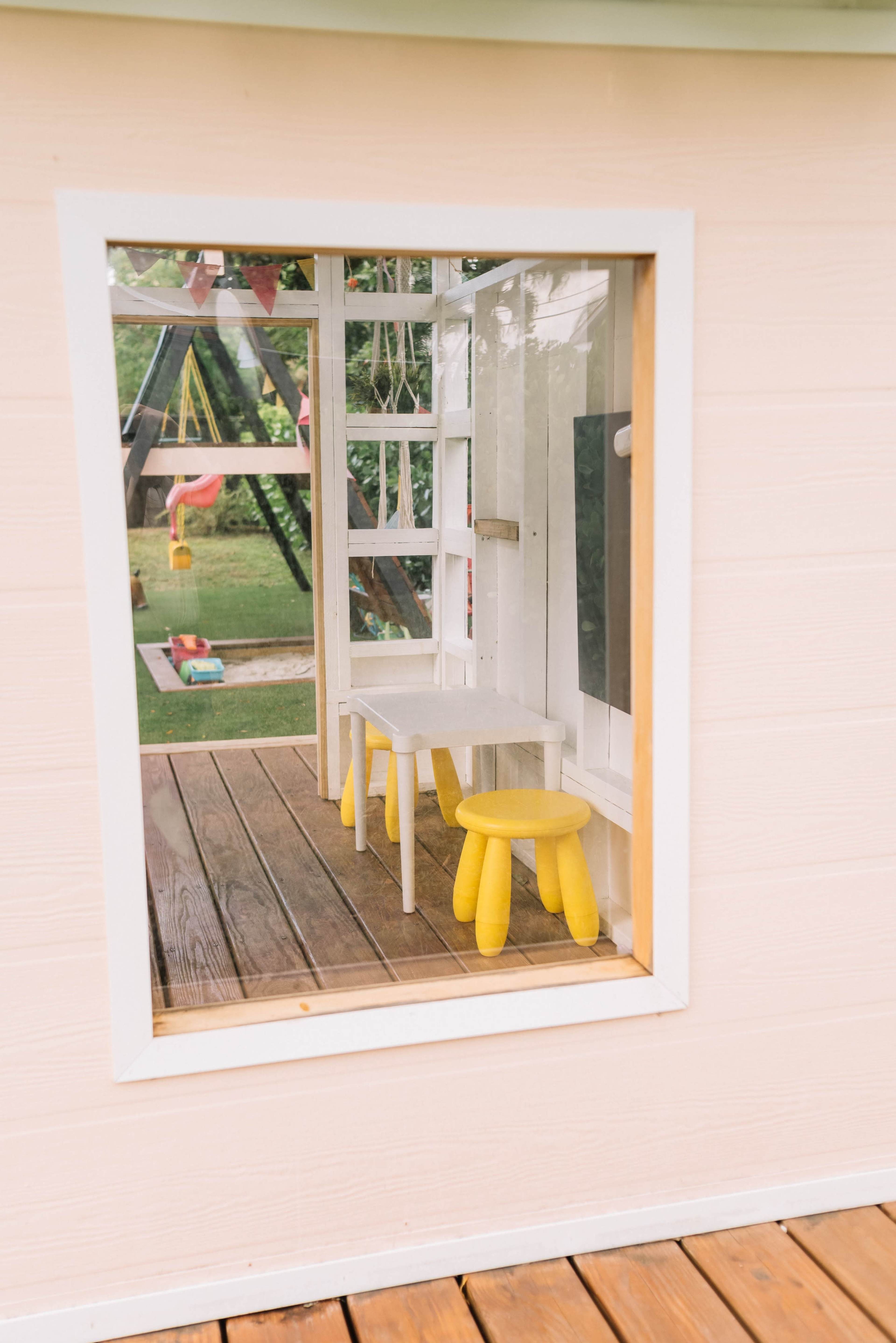 A view through a window shows a small playroom with a white table and two yellow stools, overlooking a backyard playground.