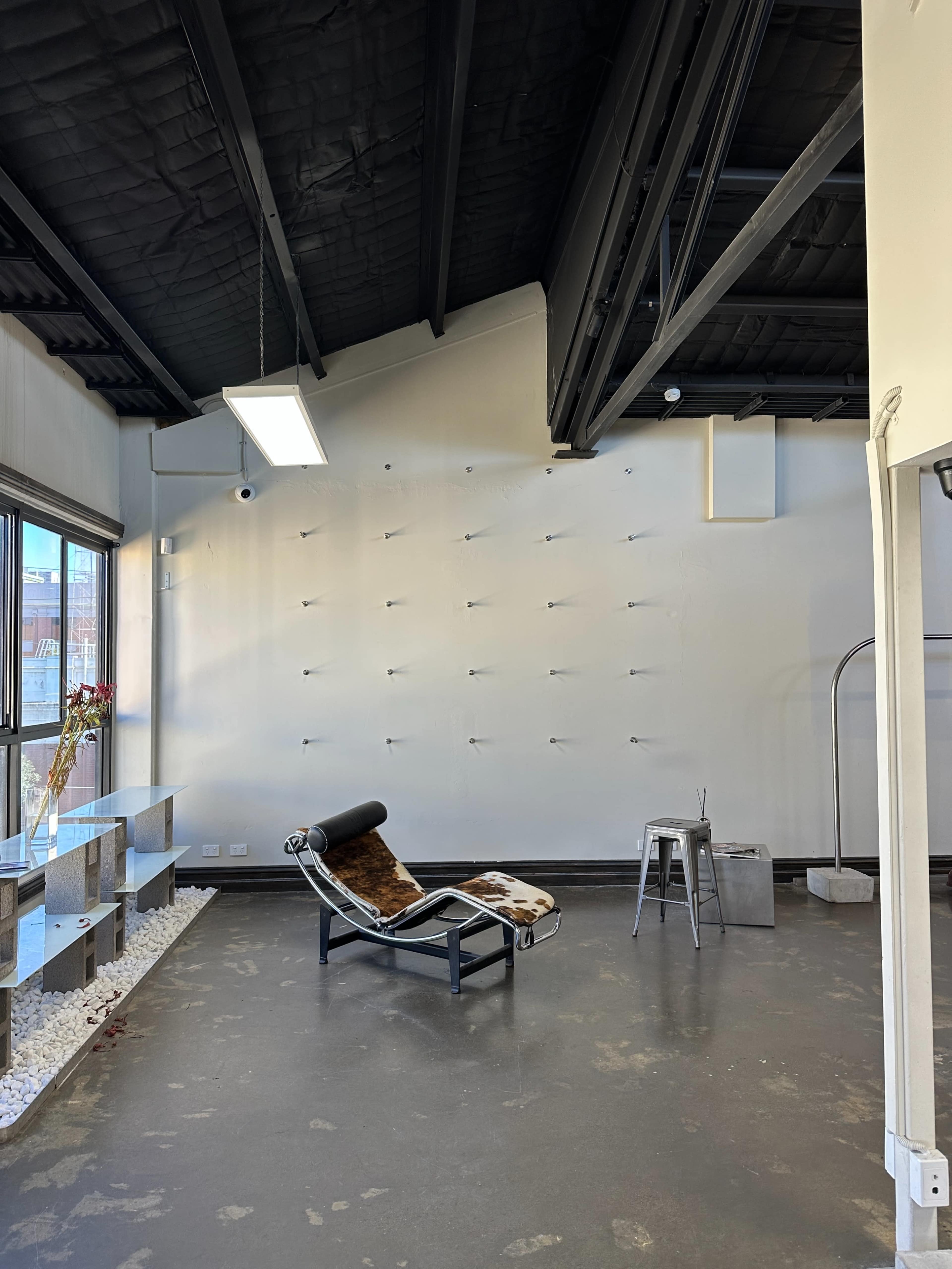 The image shows a modern, minimalist interior space featuring a lounge chair, a small metal stool, and an empty wall with multiple hanging fixtures.