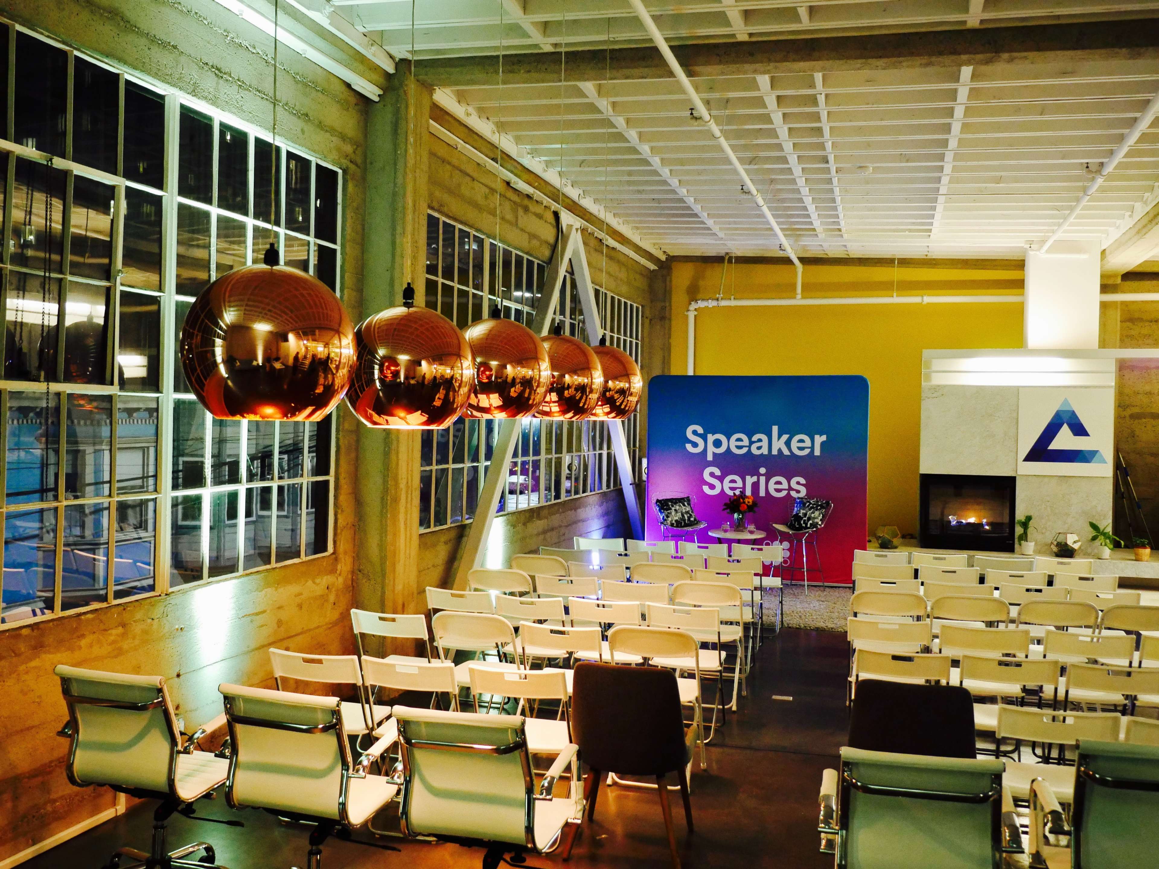 A well-lit event space features rows of chairs facing a colorful backdrop with the words "Speaker Series," alongside modern pendant lights and a fireplace.