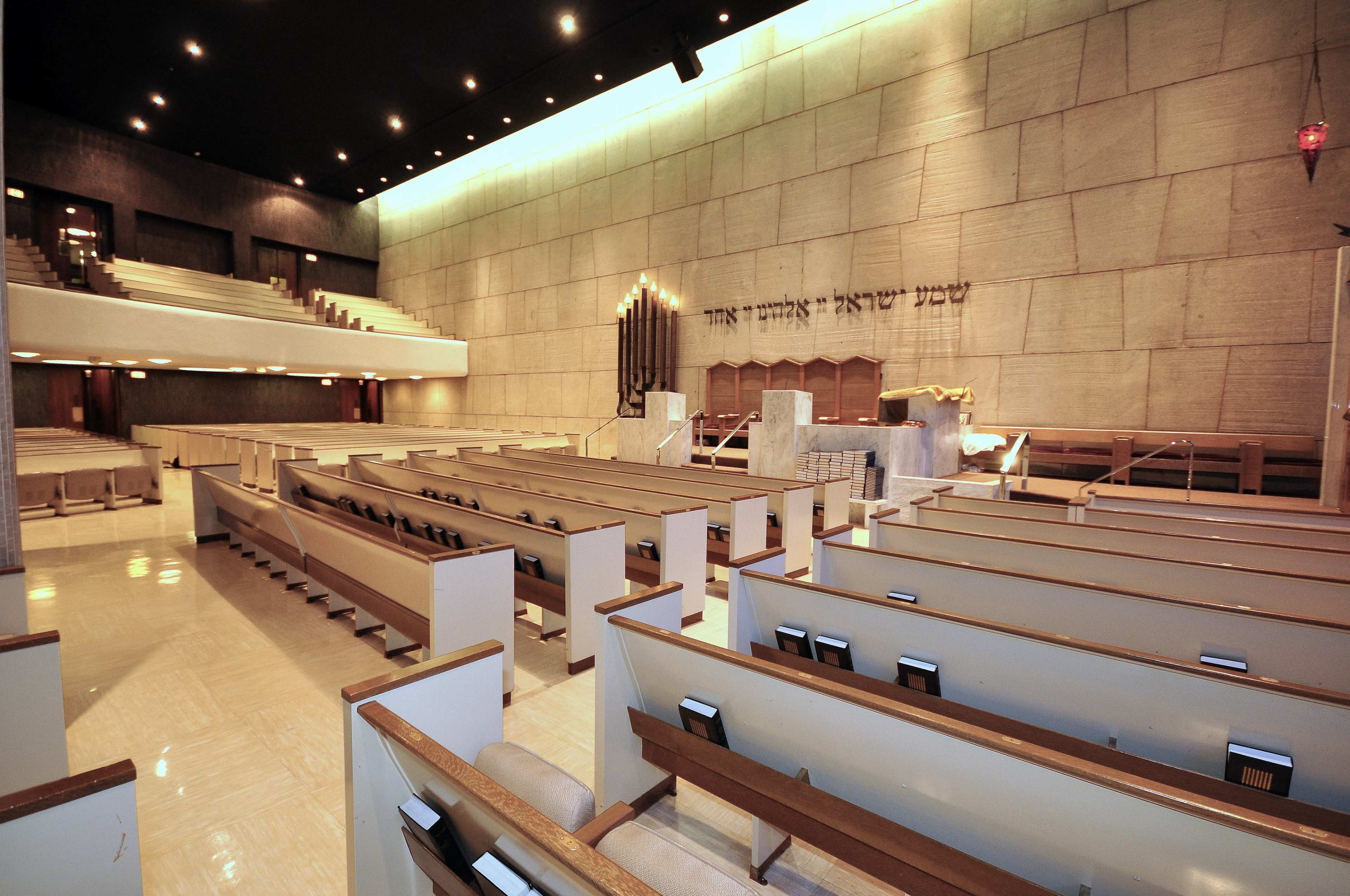The interior of a modern synagogue features rows of wooden pews facing a central bima, with a large textured wall behind it displaying Hebrew text.