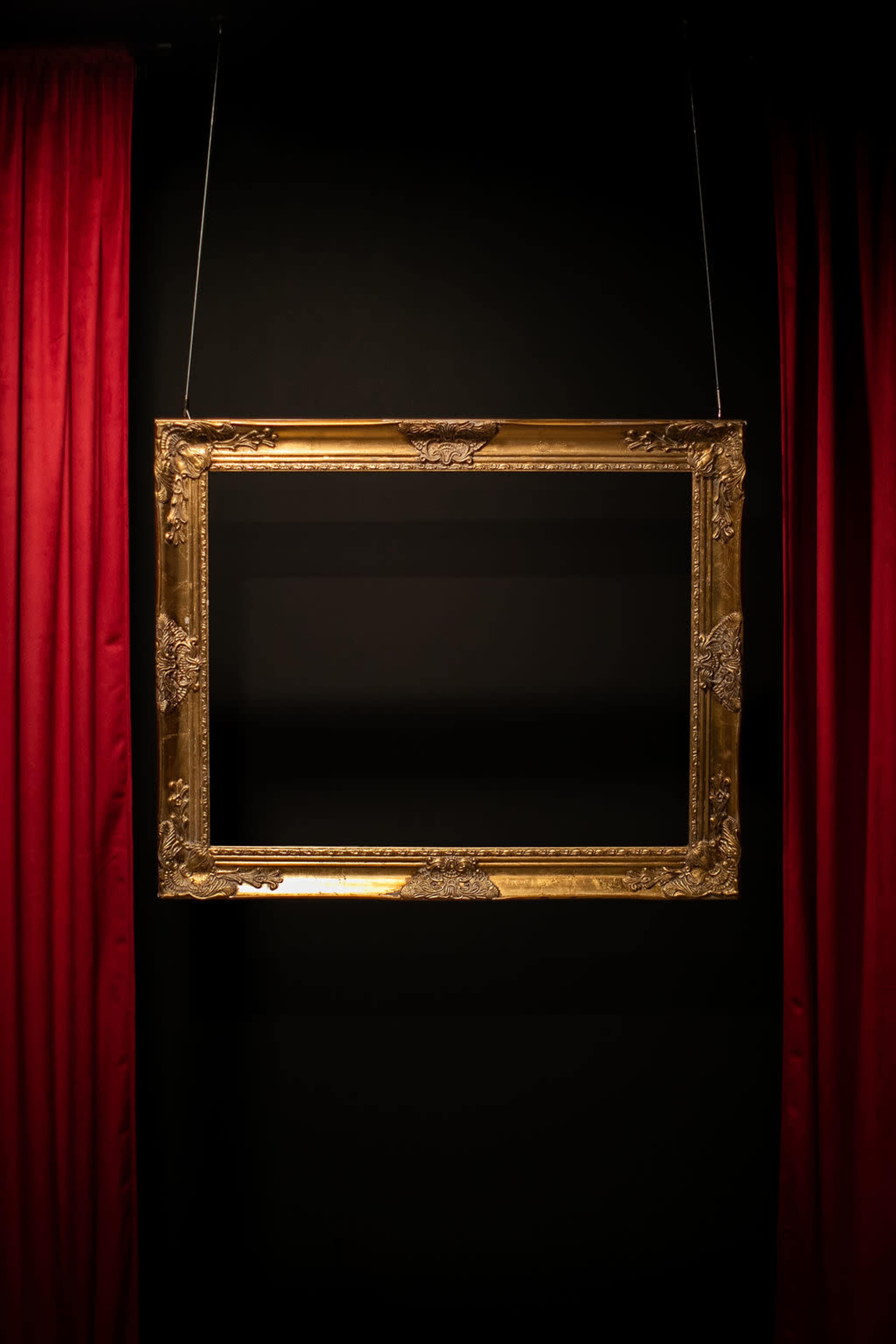 A gold, ornate picture frame hangs against a black wall, flanked by red curtains.