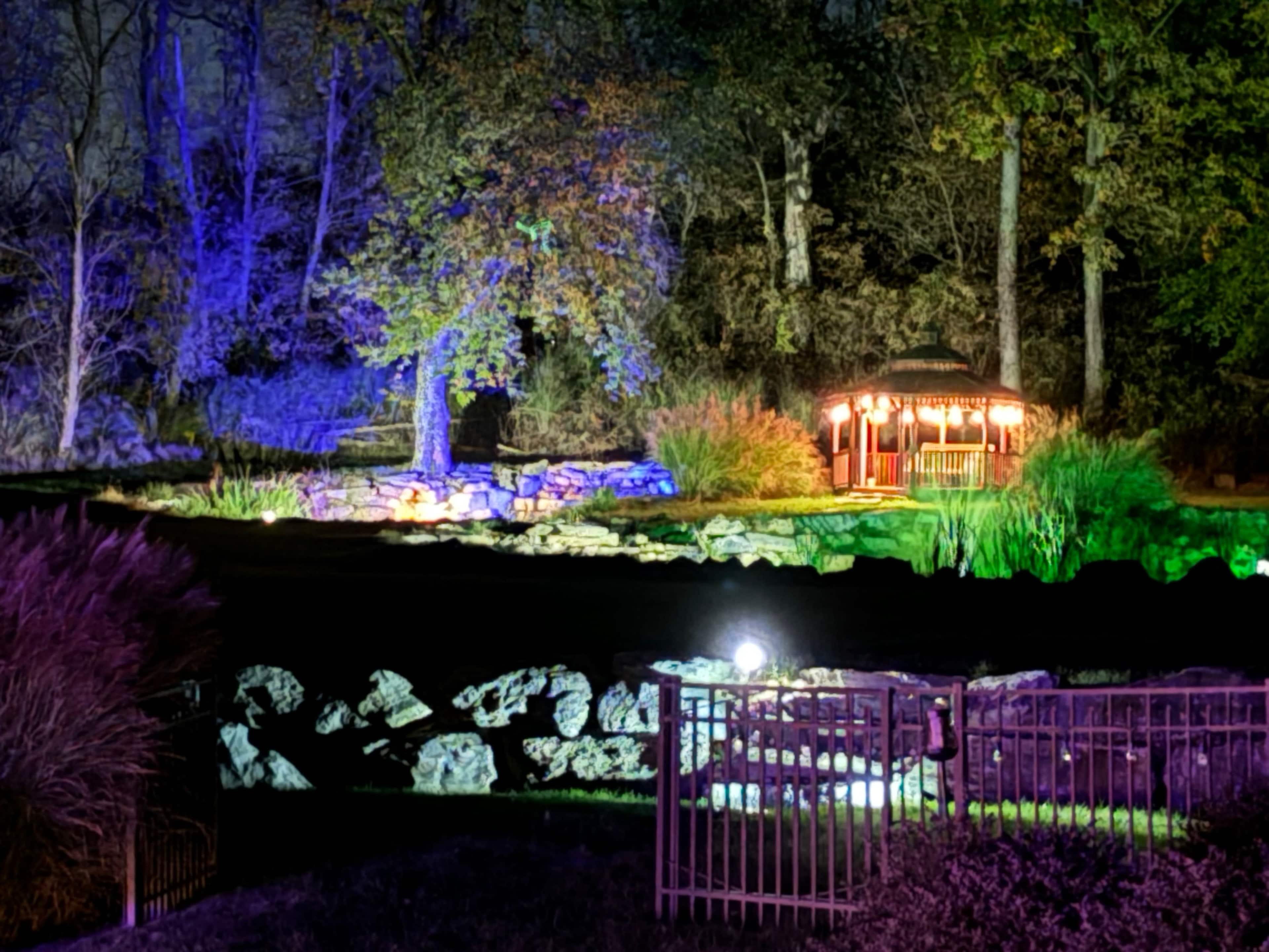 A brightly lit gazebo stands near a pond surrounded by trees, illuminated by colorful lights at night.