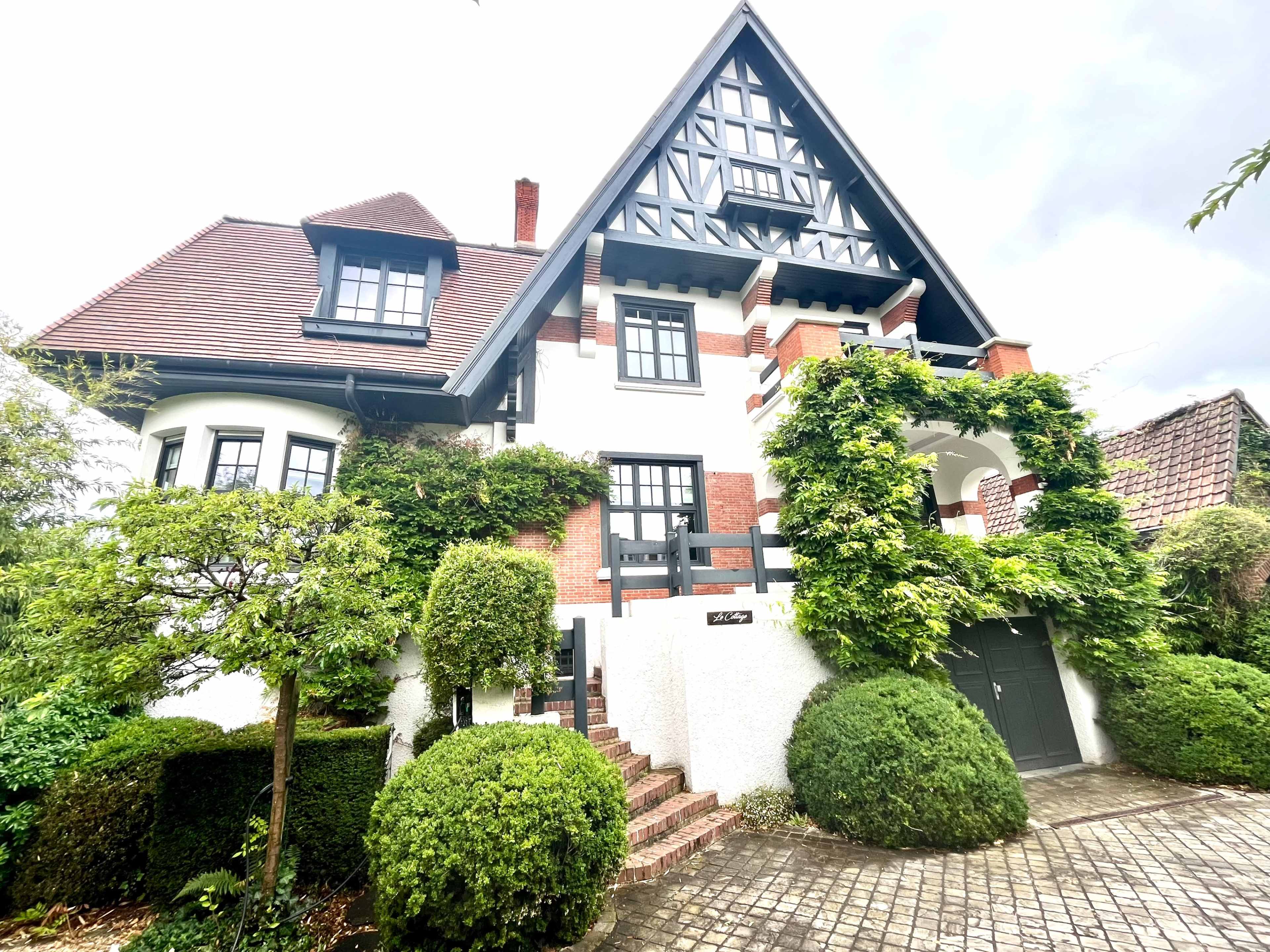 A large, three-story house features a triangular roof, decorative wooden elements, and is surrounded by neatly trimmed hedges and brick pathways.