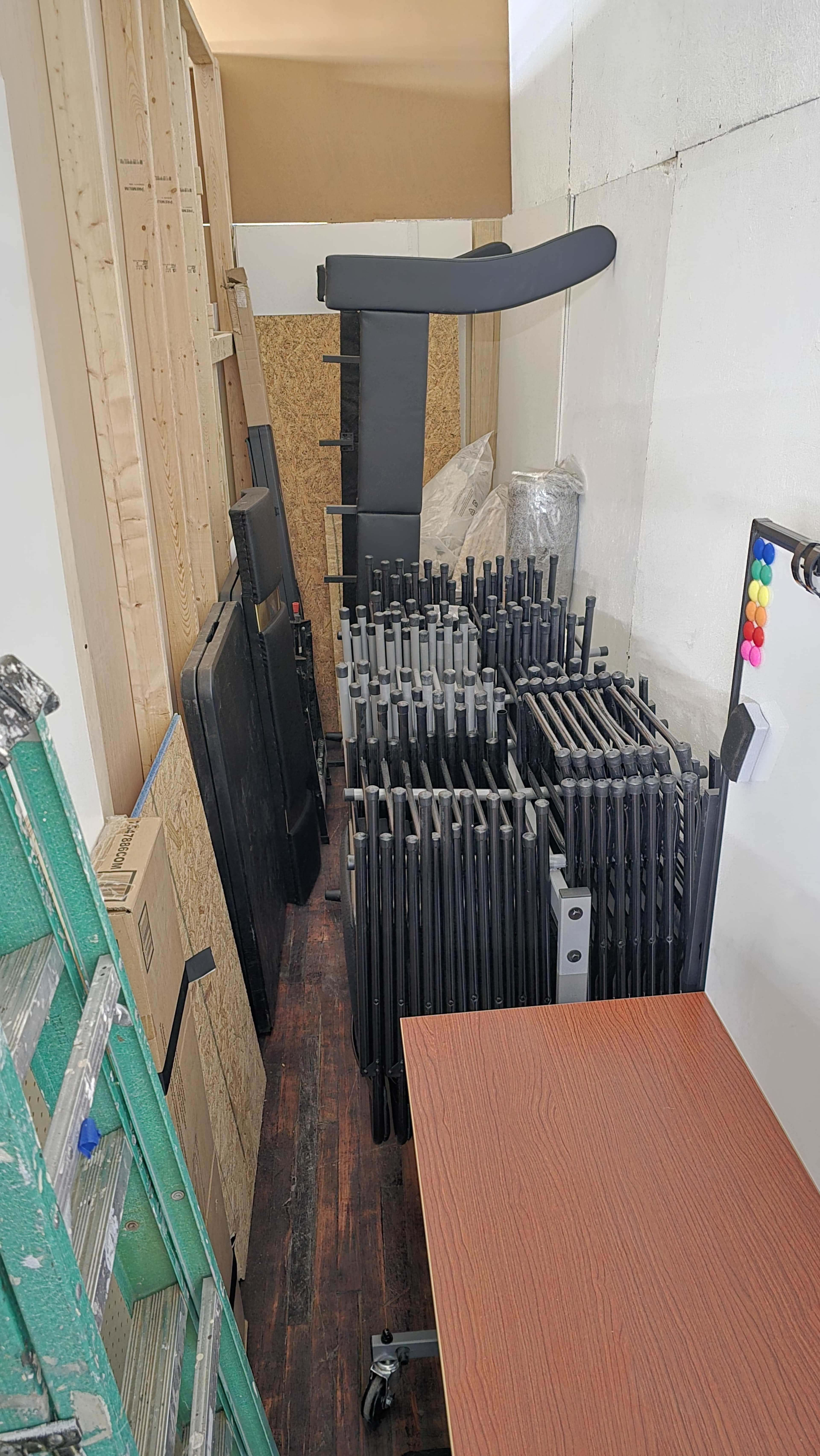 The image shows a narrow room filled with stacked black folding tables, leaning wooden panels, a green ladder, and a brown table along one side.