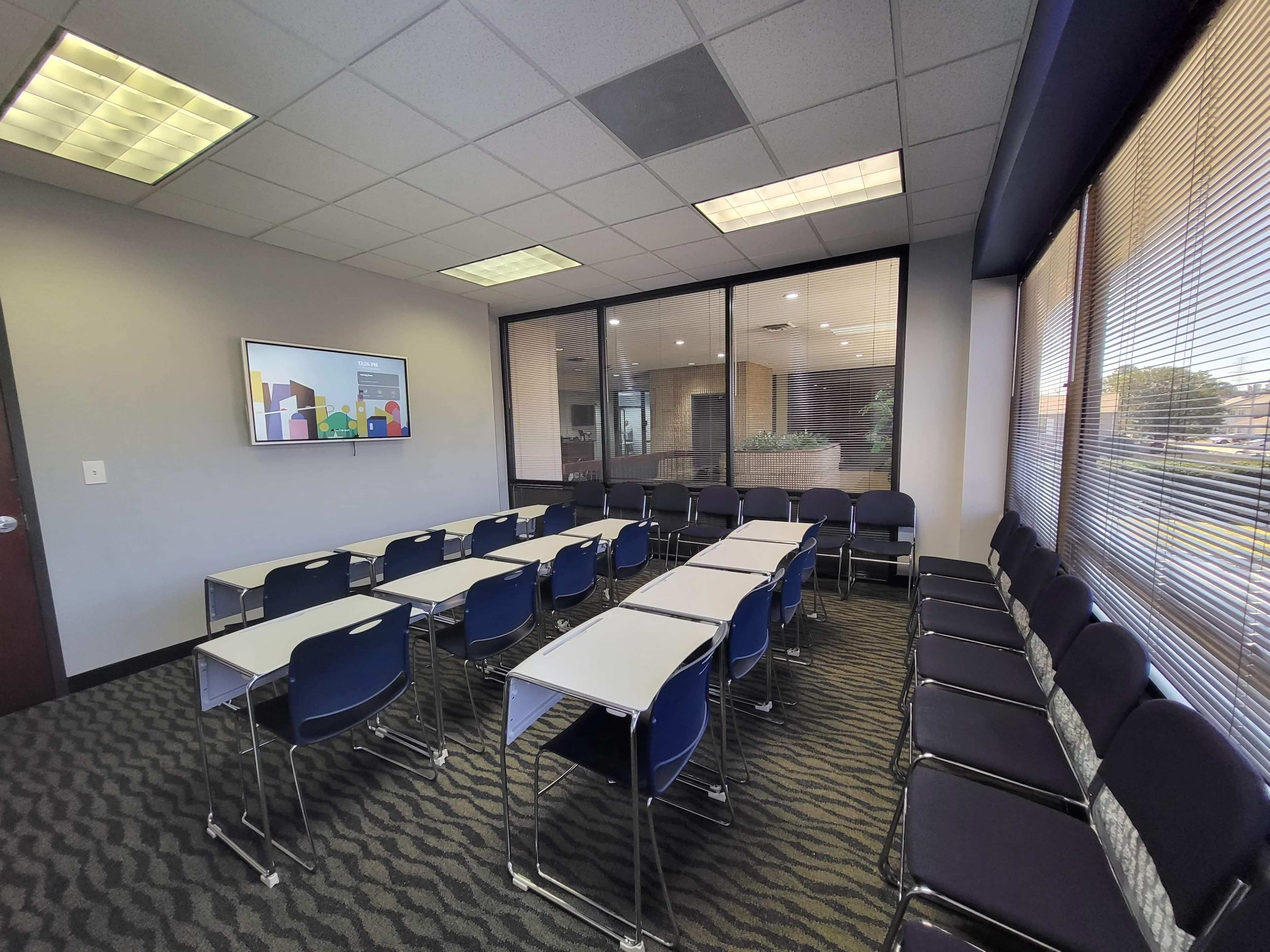 A conference room features rows of chairs and tables facing a wall-mounted screen beside large windows with a view outside.